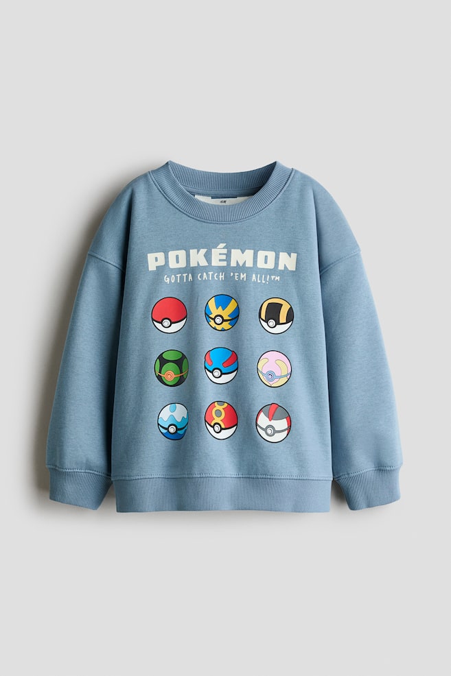 En stÃ¸vet blÃ¥ sweatshirt ses forfra og fremviser en ribbet rund halsudskÃ¦ring, sÃ¦nkede skuldre samt ribbede manchetter og sÃ¸m. PÃ¥ brystet ses et hvidt grafisk tryk med teksten "POKï¿½MON" og "GOTTA CATCH 'EM ALL!?" over et gitter af ni flerfarvede Pokï¿½ Balls. - 1