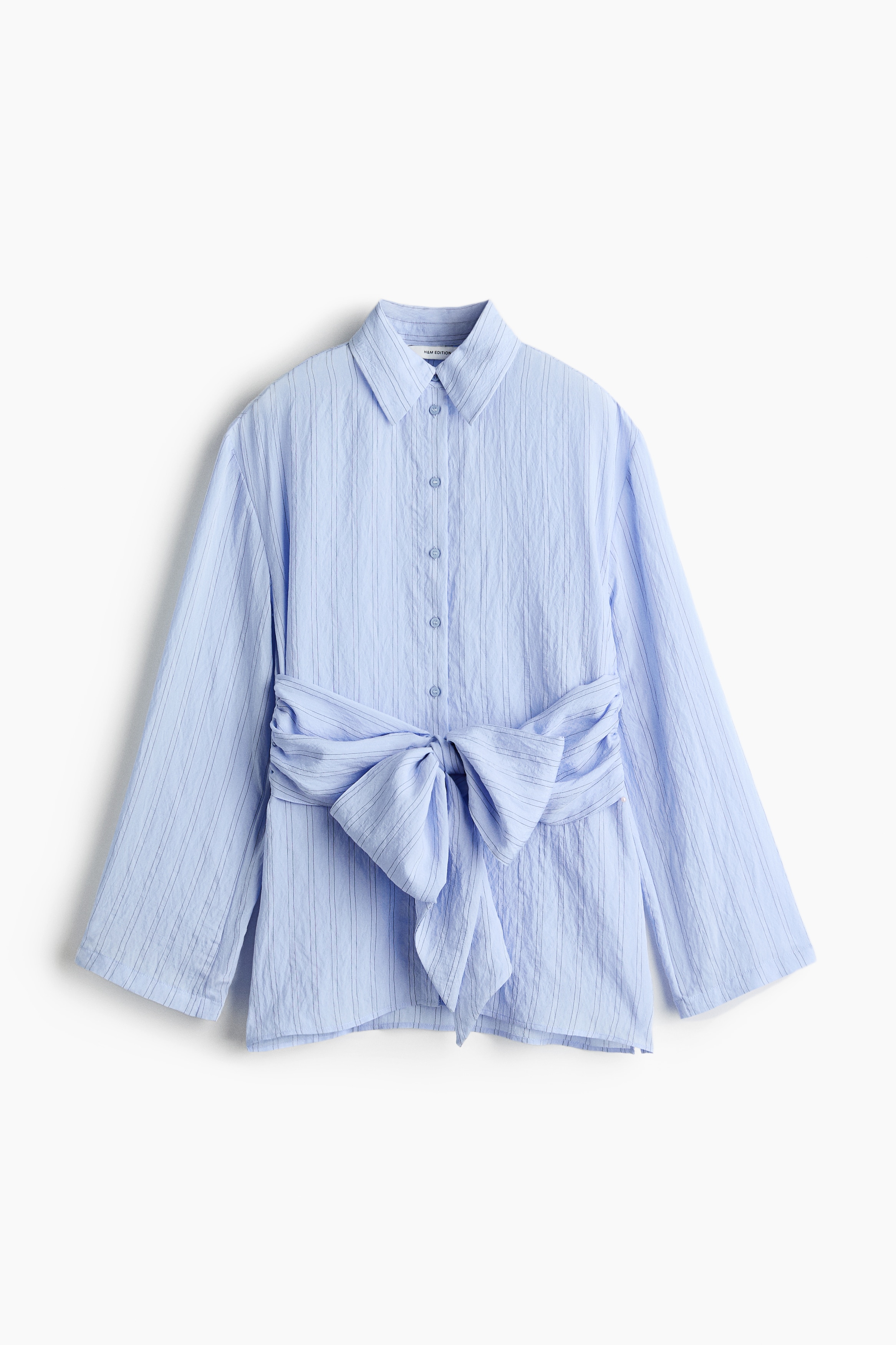 La modelo lleva una camisa de manga larga de color azul claro con botones, que presenta un patrÃ³n vertical texturizado y una faja de tela integrada anudada a la cintura. La camisa se combina con unos pantalones anchos de color azul oscuro.
