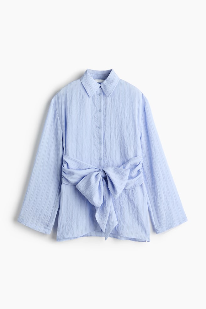 A light blue button-down shirt is shown from the front, featuring a crinkled vertical texture, a pointed collar, and wide, long sleeves. A matching fabric sash cinches the waist, tied into a large decorative bow at the centre. - 1