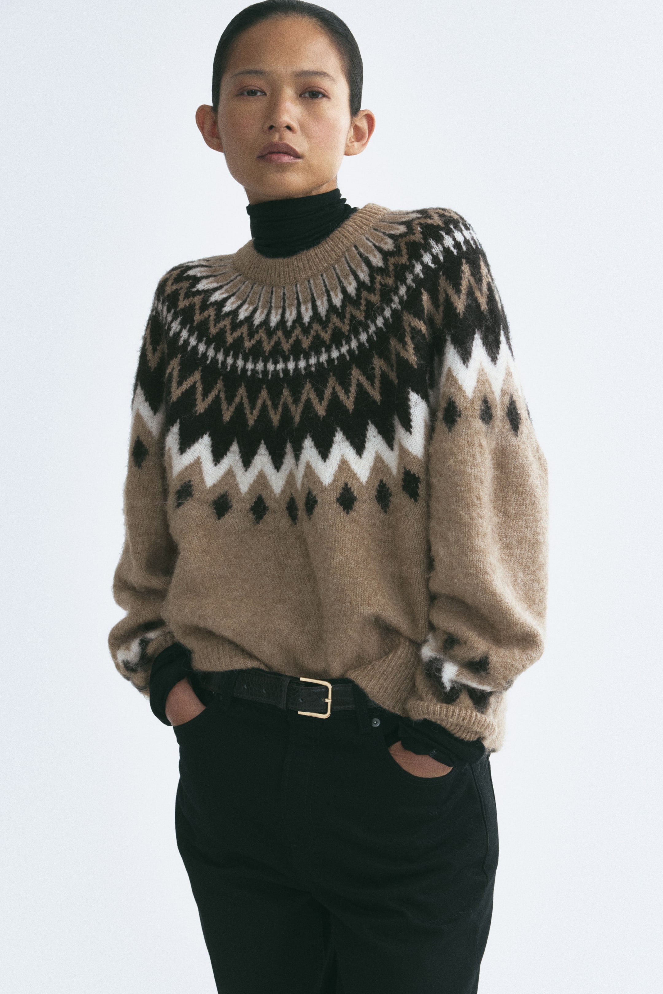 Agrandir l'image: Un pull oversize en maille jacquard avec une finition brossée et un motif géométrique beige, noir et blanc est porté par le mannequin par-dessus un col roulé noir. Le pull présente un col rond bordé de côtes, de longues manches raglan ainsi que des poignets et un ourlet côtelés, et est associé à un pantalon noir.