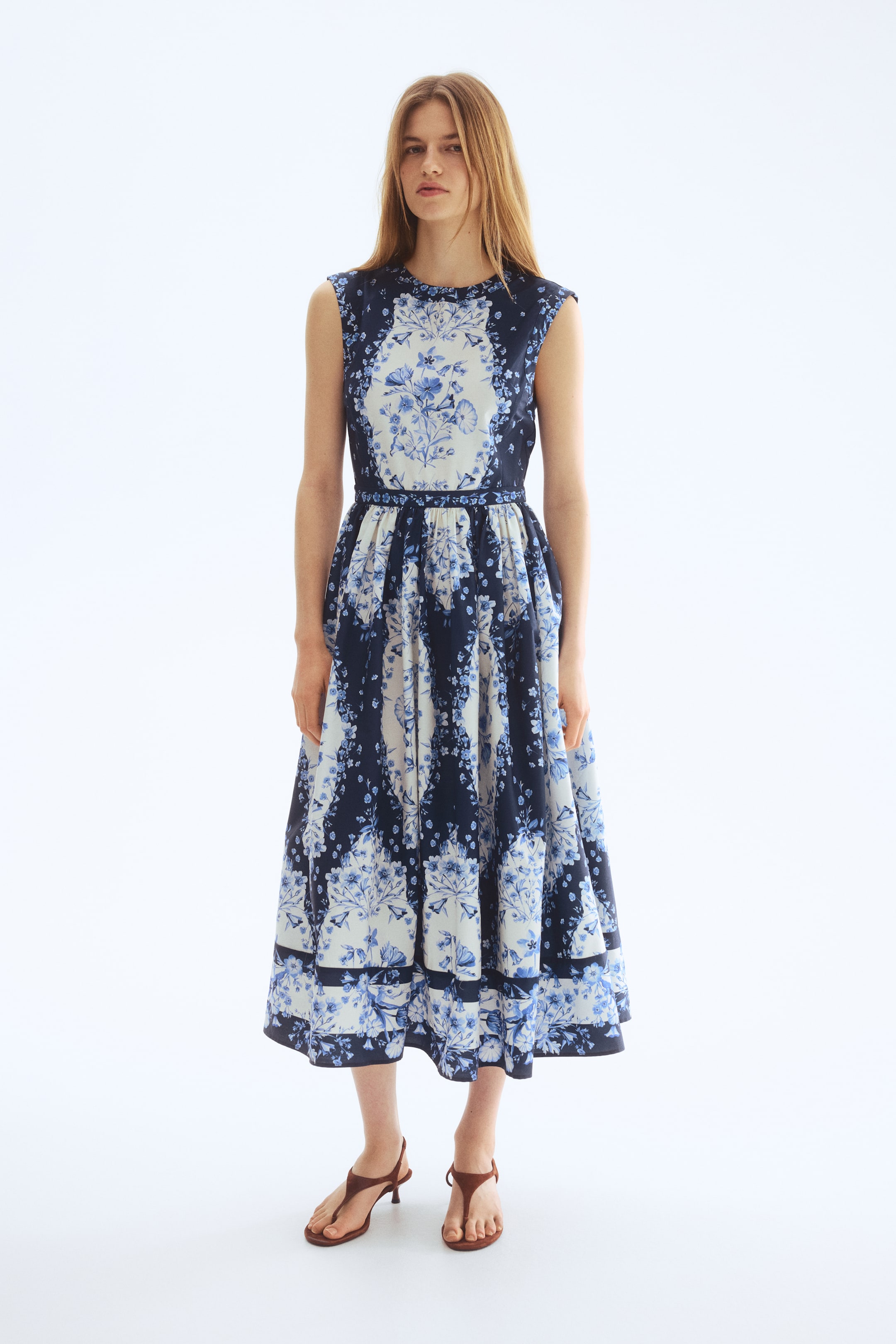 View larger image: The dress showcases a blue and white floral pattern, with a round neckline and sleeveless style. It features a fitted bodice highlighted by a distinct white central panel, complemented by a full, midi-length skirt made up of alternating patterned vertical panels and a border print near the hem.