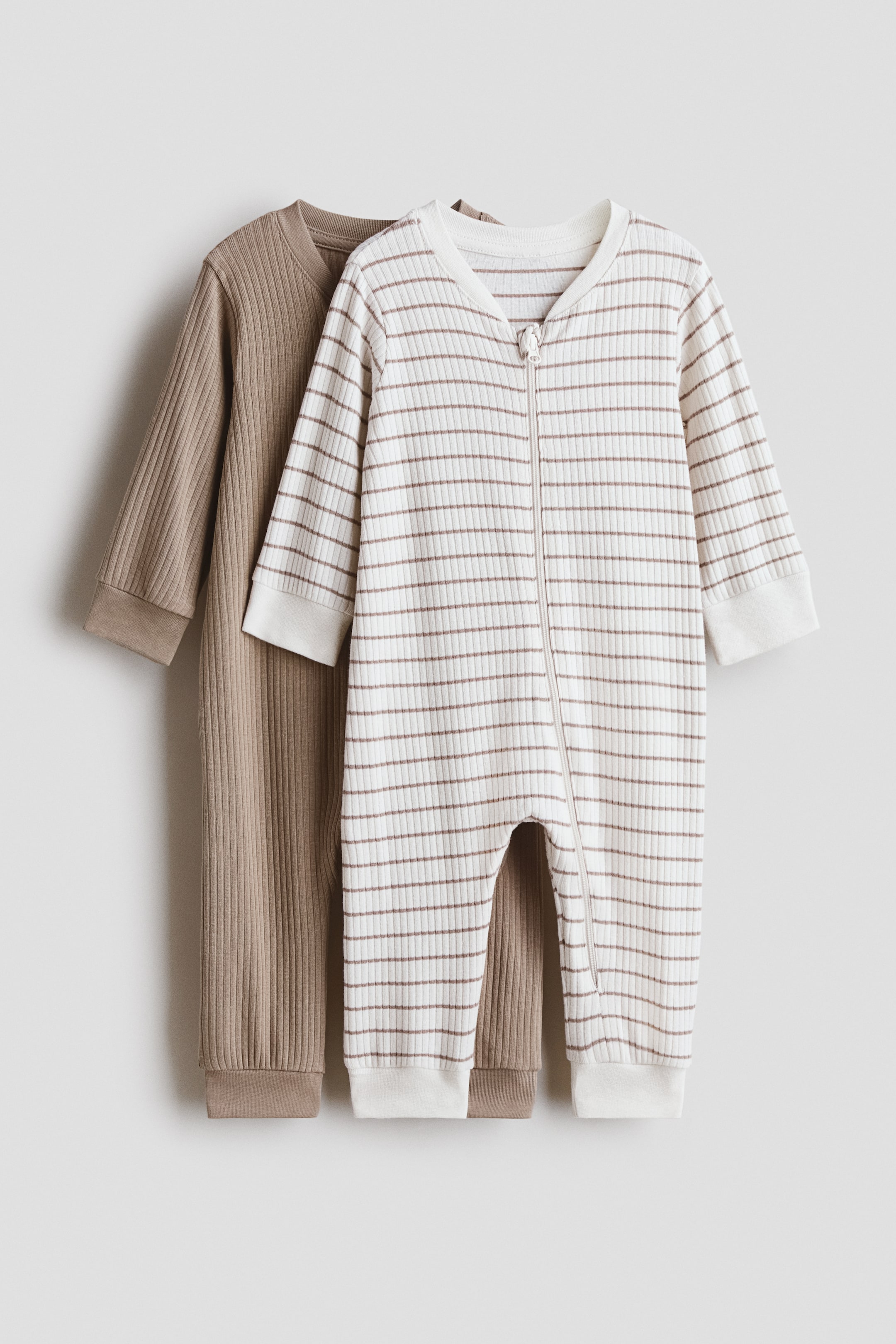 View larger image: Two ribbed knit baby rompers are hanging, showcasing their front. The front romper is off-white with thin brown horizontal stripes, featuring a full-length front zipper, ribbed cuffs, and ribbed ankles. The romper behind it is a solid light brown, also featuring ribbed cuffs and ribbed ankles.