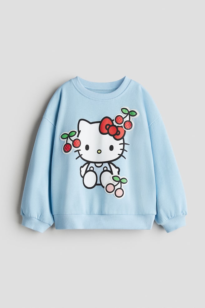 Jasnoniebieska bluza prezentuje swój przedni widok, z prążkowanym dekoltem, prążkowanymi mankietami i prążkowanym dołem. Duża grafika Hello Kitty jest umieszczona na środku klatki piersiowej, otoczona wieloma aplikacjami wiśni. - 1