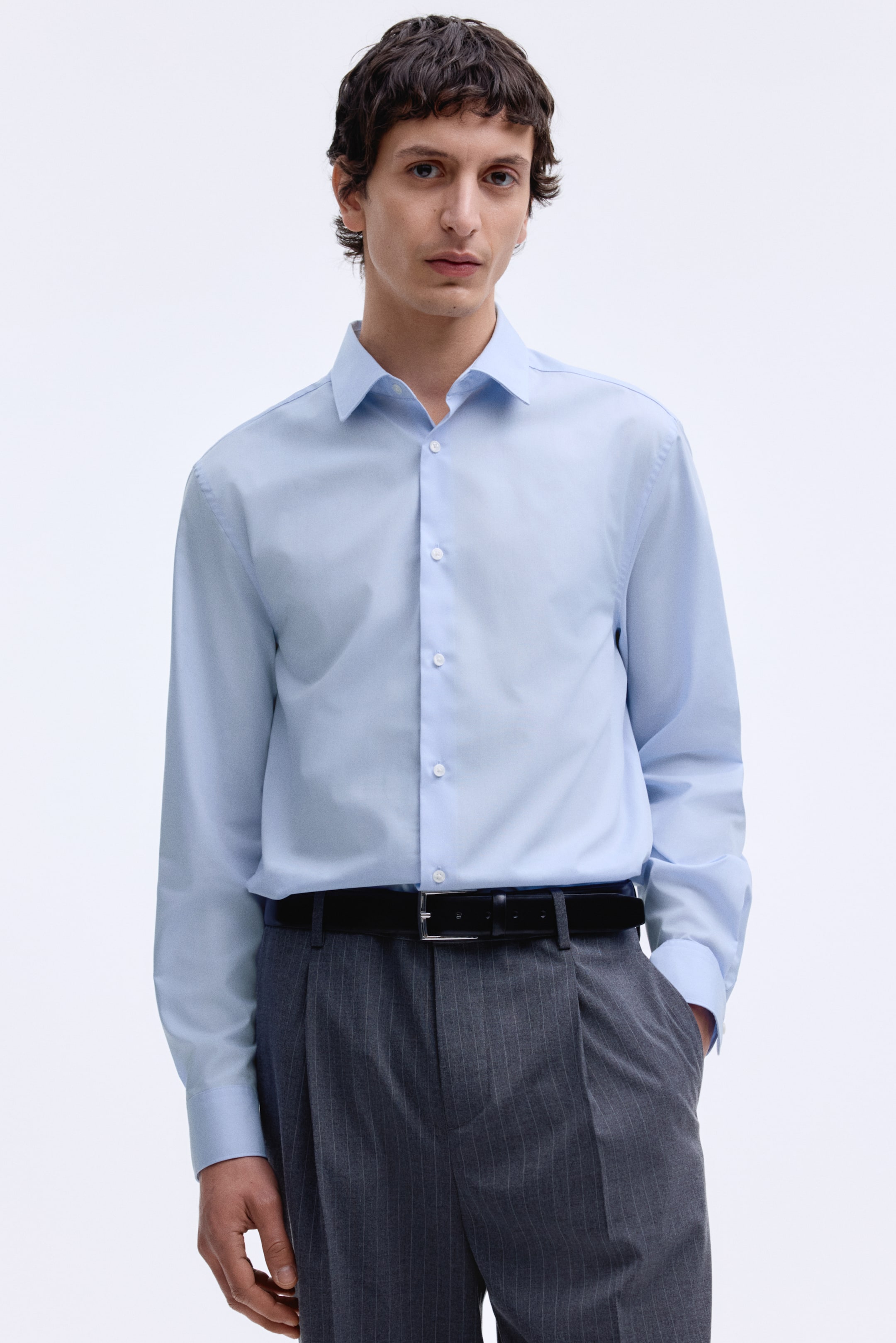 View larger image: A light blue dress shirt is showcasing its front, featuring a classic collar and white buttons running down the placket. Its long sleeves extend to buttoned cuffs, and it is worn tucked into trousers and secured by a belt.