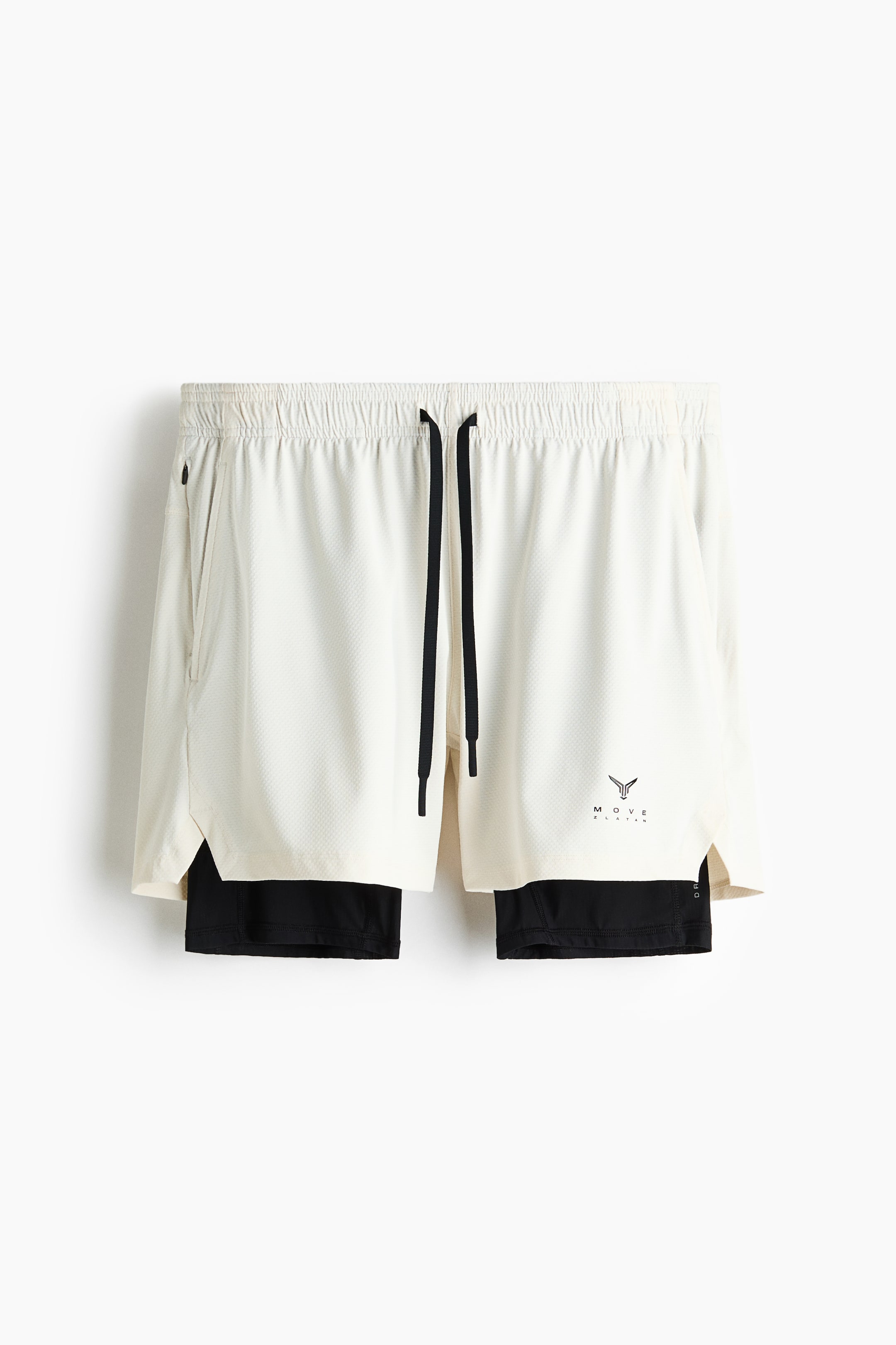 Ampliar la imagen: Un par de shorts atléticos dos en uno está de frente, con una capa exterior de color blanco roto, con textura de malla, con una pretina elástica, cordones negros, un bolsillo con cremallera en el lado izquierdo y un logotipo negro en el muslo derecho. Una capa interior negra se extiende por debajo de los shorts exteriores.