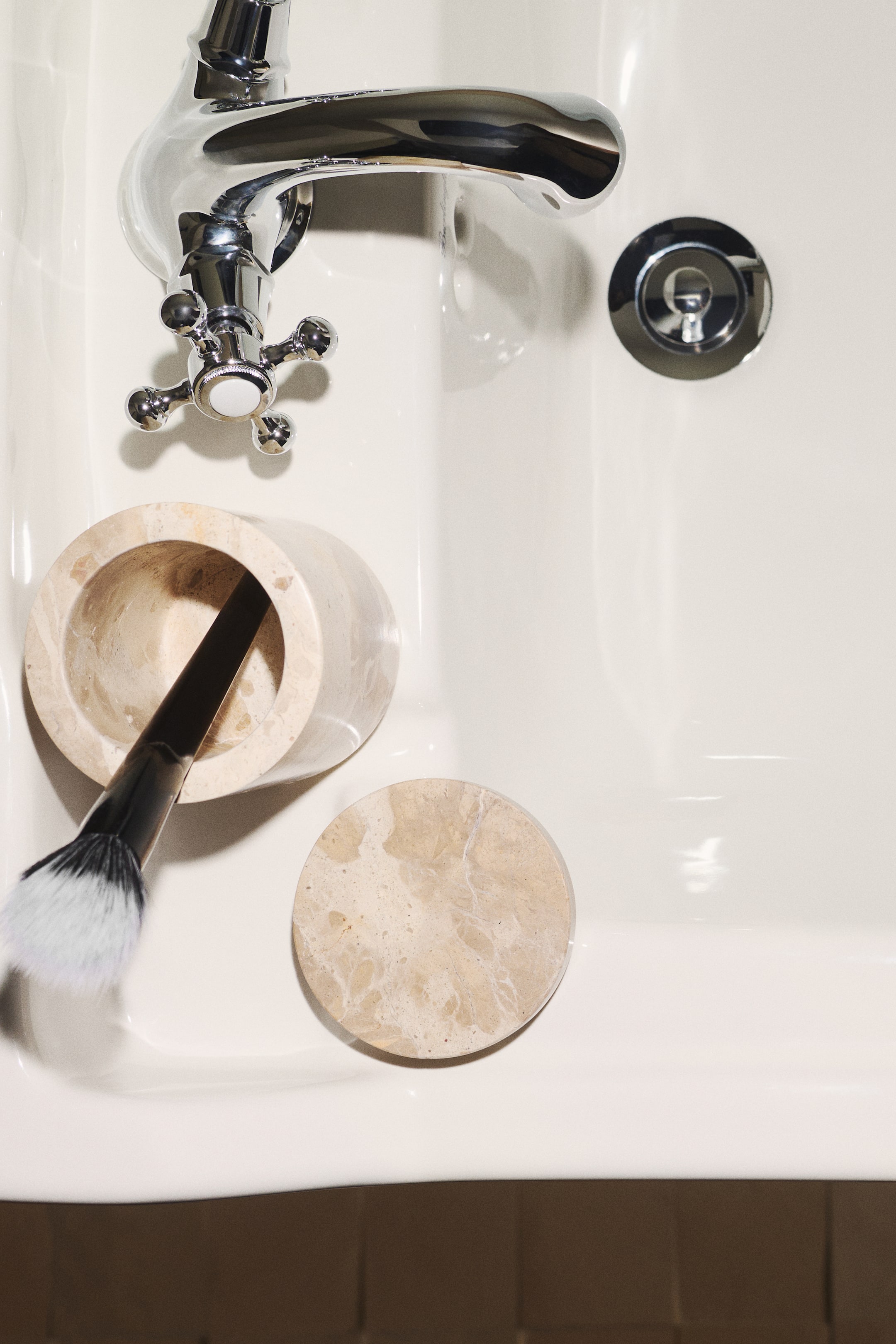 View larger image: A cylindrical lidded jar in beige marble is sitting open in a top-down view, with its separate circular lid lying beside it. A makeup brush rests inside the open jar, showing the natural veining and light tan colour variations of the marble.