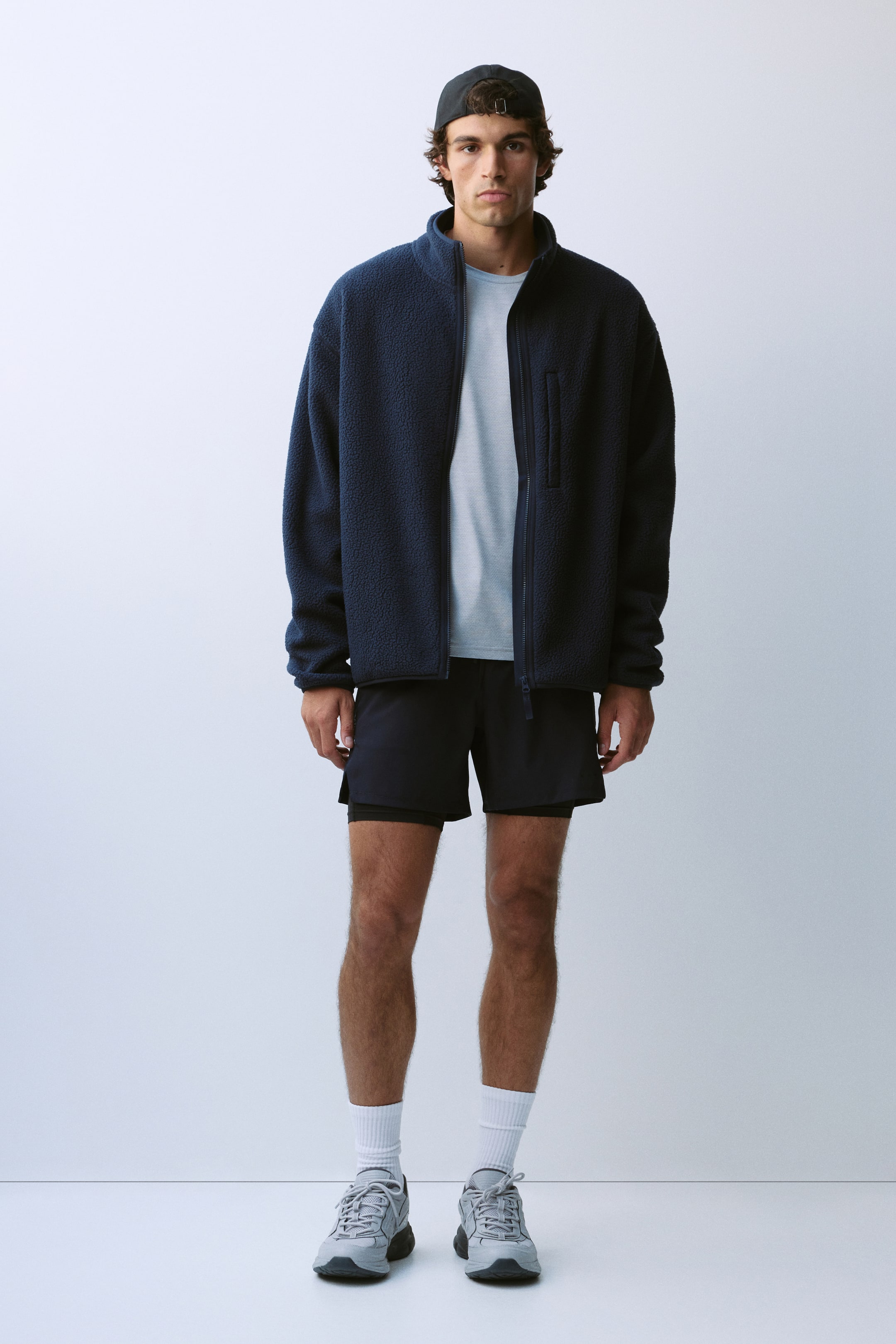 View larger image: The model is wearing a dark blue teddy fleece activewear jacket featuring a high stand collar, a front zipper left open, and a vertical zippered chest pocket. The jacket is layered over a light grey T-shirt and paired with dark athletic shorts.
