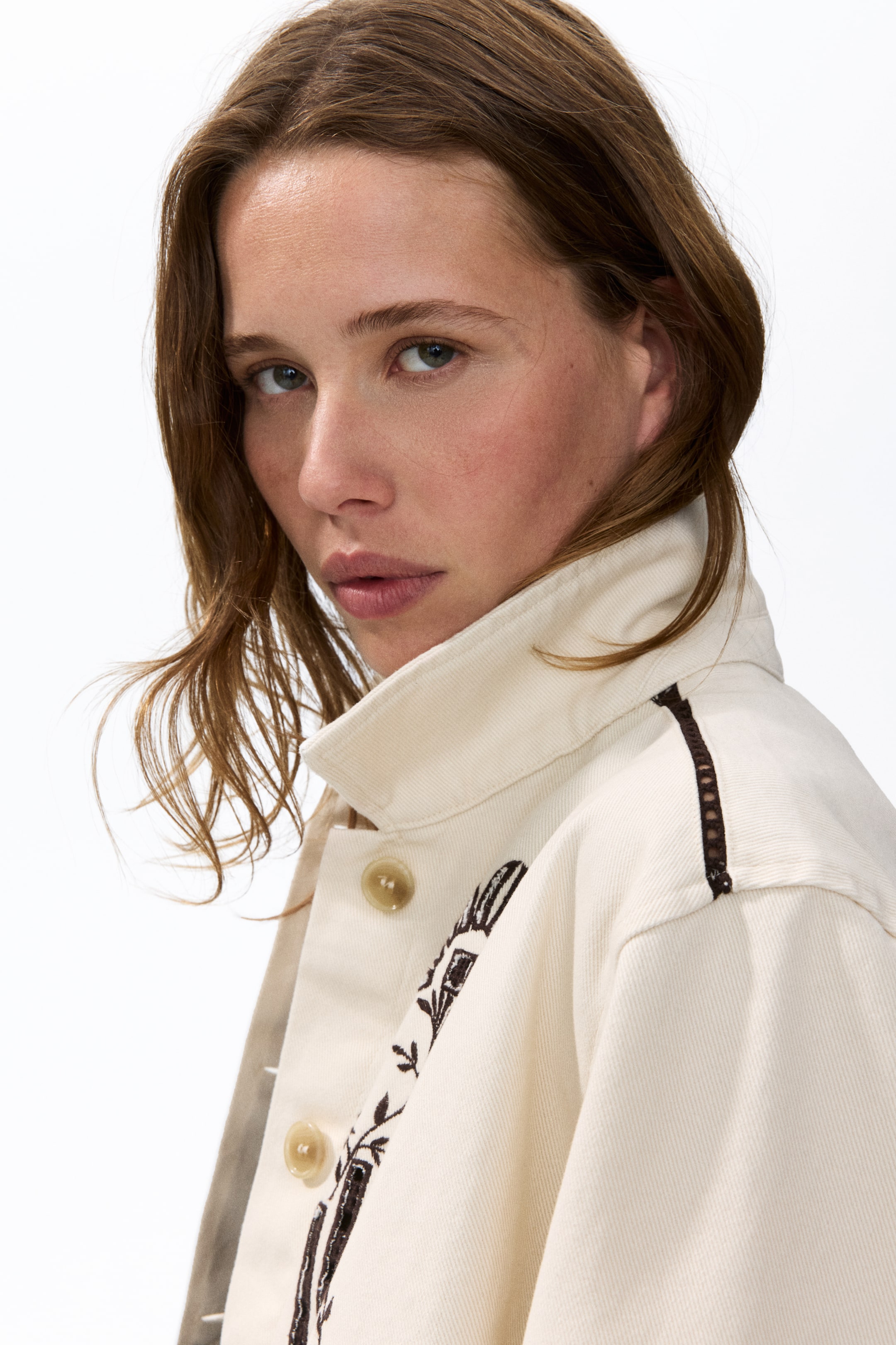 View larger image: A cream-coloured jacket featuring a standing pointed collar and two visible light brown buttons on the front placket, with dark brown embroidery depicting a pattern vertically along the front and a dark brown openwork trim running along the shoulder seam.