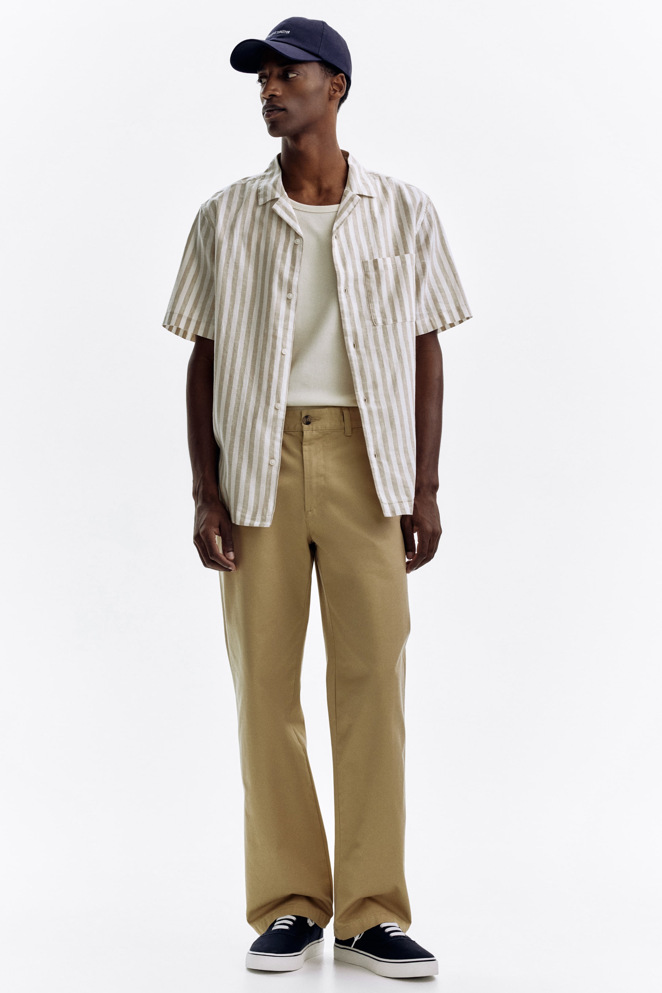 Grösseres Bild ansehen: Ein kurzärmeliges, vertikal gestreiftes Hemd in Beige und Weiß mit einem Camp-Kragen und einer linken Brusttasche wird offen über einem schlichten cremefarbenen T-Shirt getragen. Dieses Ensemble wird mit locker sitzenden khakifarbenen Hosen, dunkelblauen Canvas-Sneakers mit weißen Schnürsenkeln und einer dunkelblauen Baseballkappe mit weißer Stickerei vervollständigt.