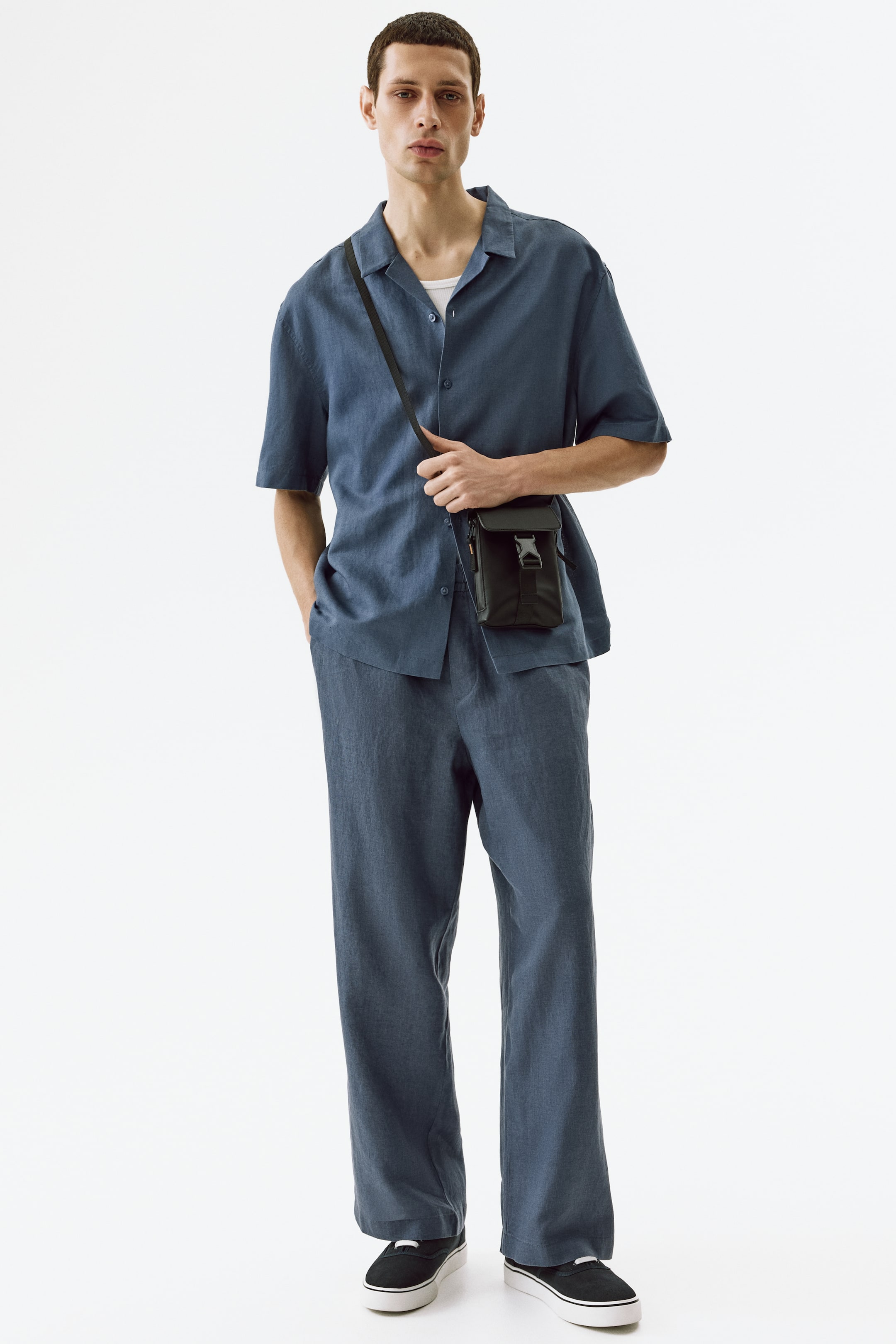 Grotere afbeelding bekijken: Een bijpassend blauw linnen kampkraagshirt met korte mouwen en wijde broek worden gepresenteerd. Het shirt met een ontspannen pasvorm heeft zichtbare knopen aan de voorkant, een wit onderhemd dat eronder zichtbaar is, en wordt los gedragen, terwijl de wijde broek recht naar de enkels valt.