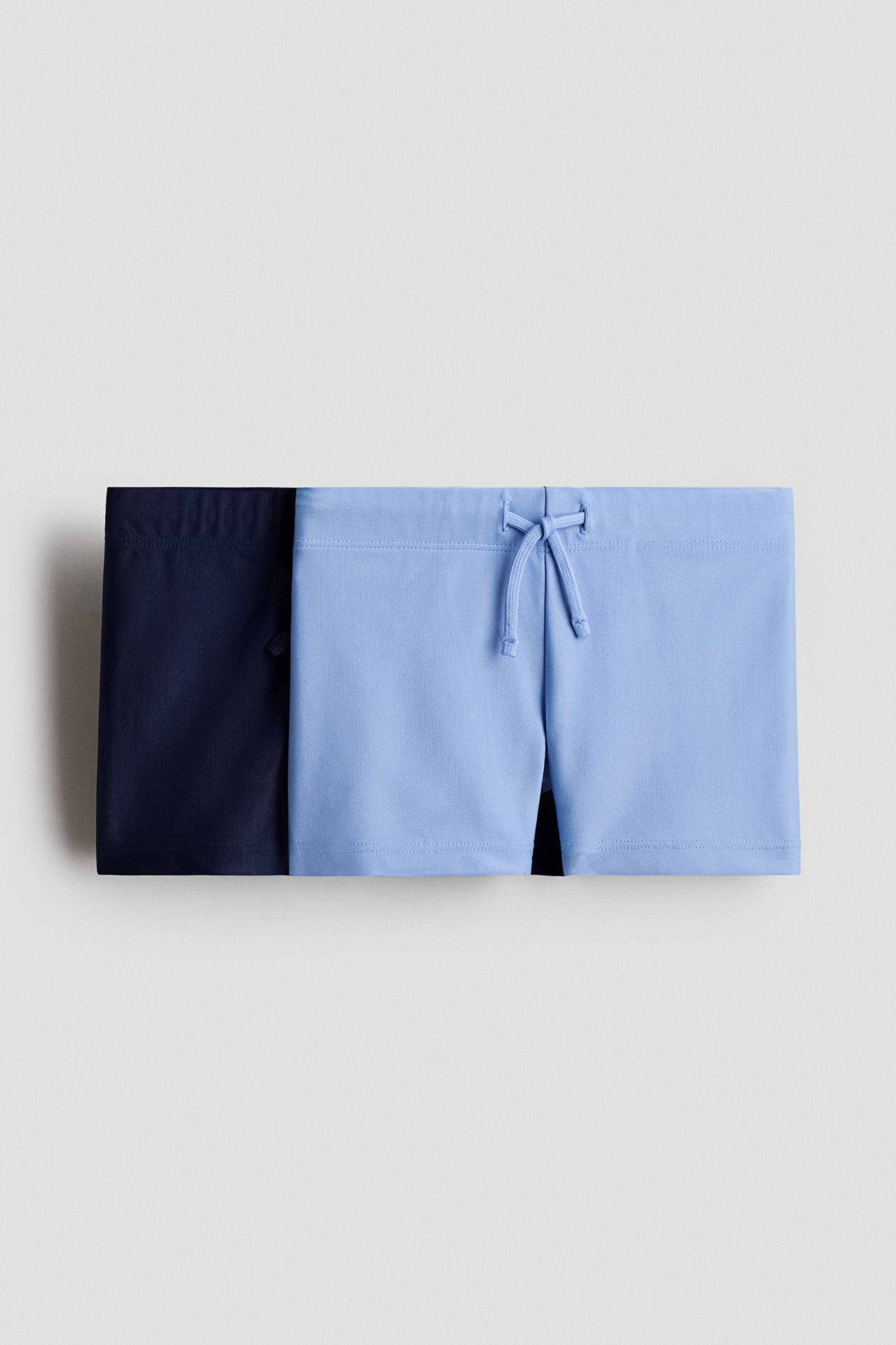 View larger image: "Two pairs of shorts showcase their design, with a light blue pair positioned in front of a navy pair. The light blue shorts feature a drawstring tied in a bow at the center front waistband."