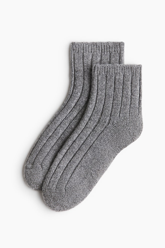 Un par de calcetines de punto acanalado en gris jaspeado estÃ¡ superpuesto mientras yace extendido, mostrando una textura gruesa con un acanalado vertical prominente que va desde los puÃ±os acanalados hasta la puntera. - 1