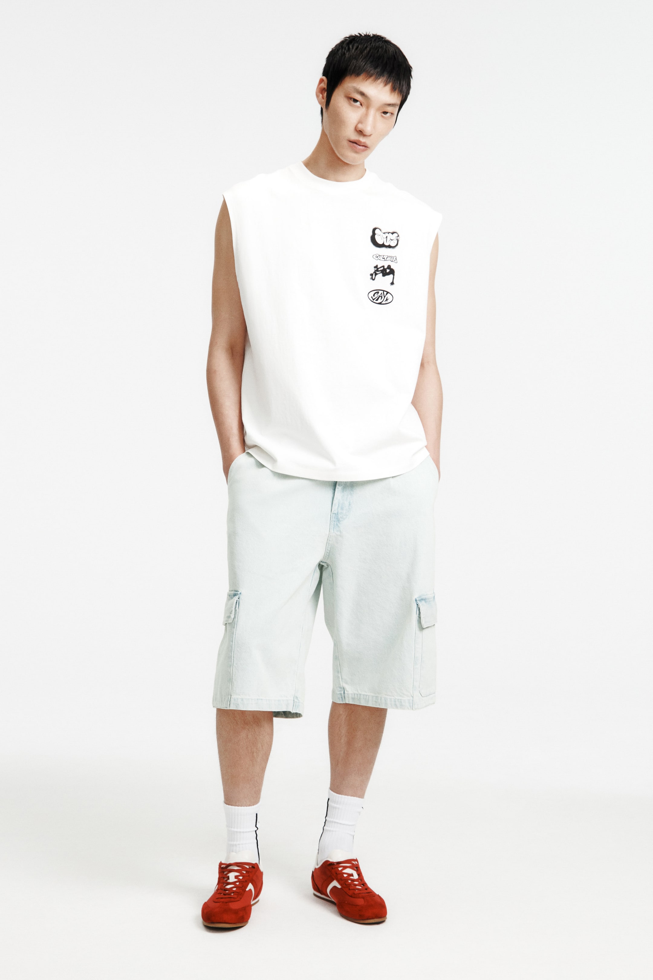 拡大画像を表示: モデルは、斜めのサイドポケットに手を入れた状態で、ライトブルーのリジッドデニムショーツを着用しています。このショーツは、トップにプリーツがあり、フラップ付きのパッチレッグポケットが特徴で、ゆったりとしたフィット感を演出しています。