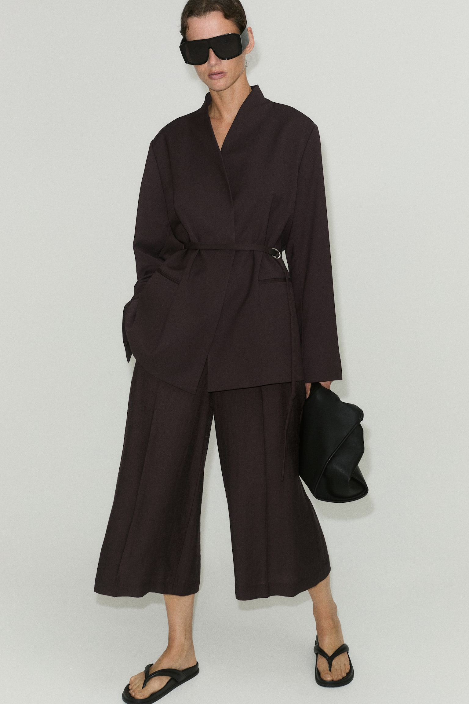 An oversized dark brown blazer is shown from the front, featuring structured shoulders, a stand collar, and a wrap-front closure secured by a narrow matching belt at the waist. The design includes subtle jetted front pockets and long, wide sleeves. - 1