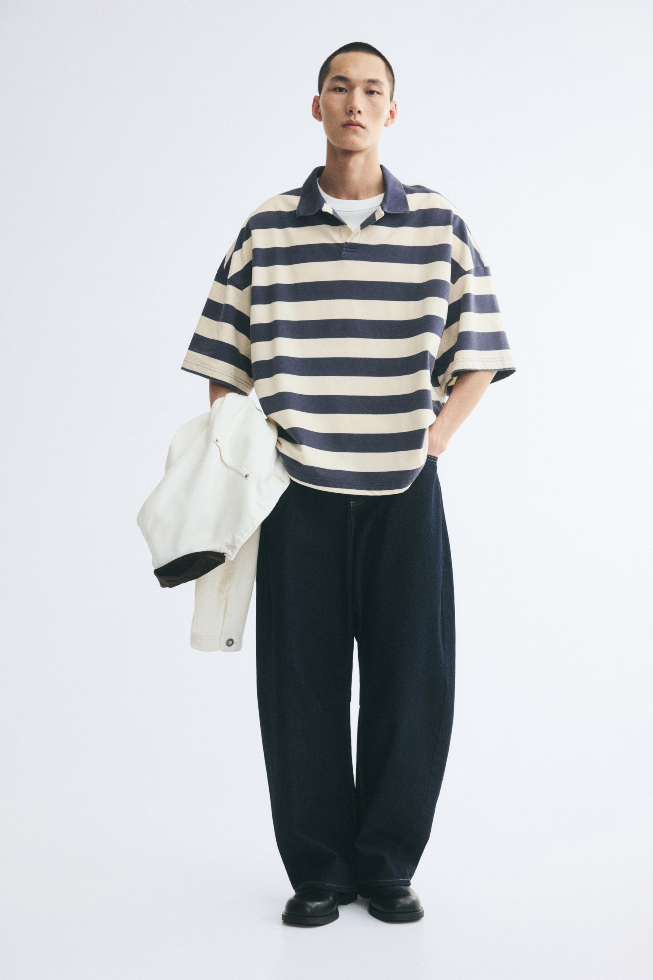 Grotere afbeelding bekijken: Een oversized poloshirt met korte mouwen en horizontale strepen in blauw en crème wordt gedragen door het model. Het shirt, ontworpen met een open kraag en verlaagde schouders, wordt gecombineerd met een wit T-shirt en donkere wijde broek, terwijl een witte jas in de hand wordt gehouden.
