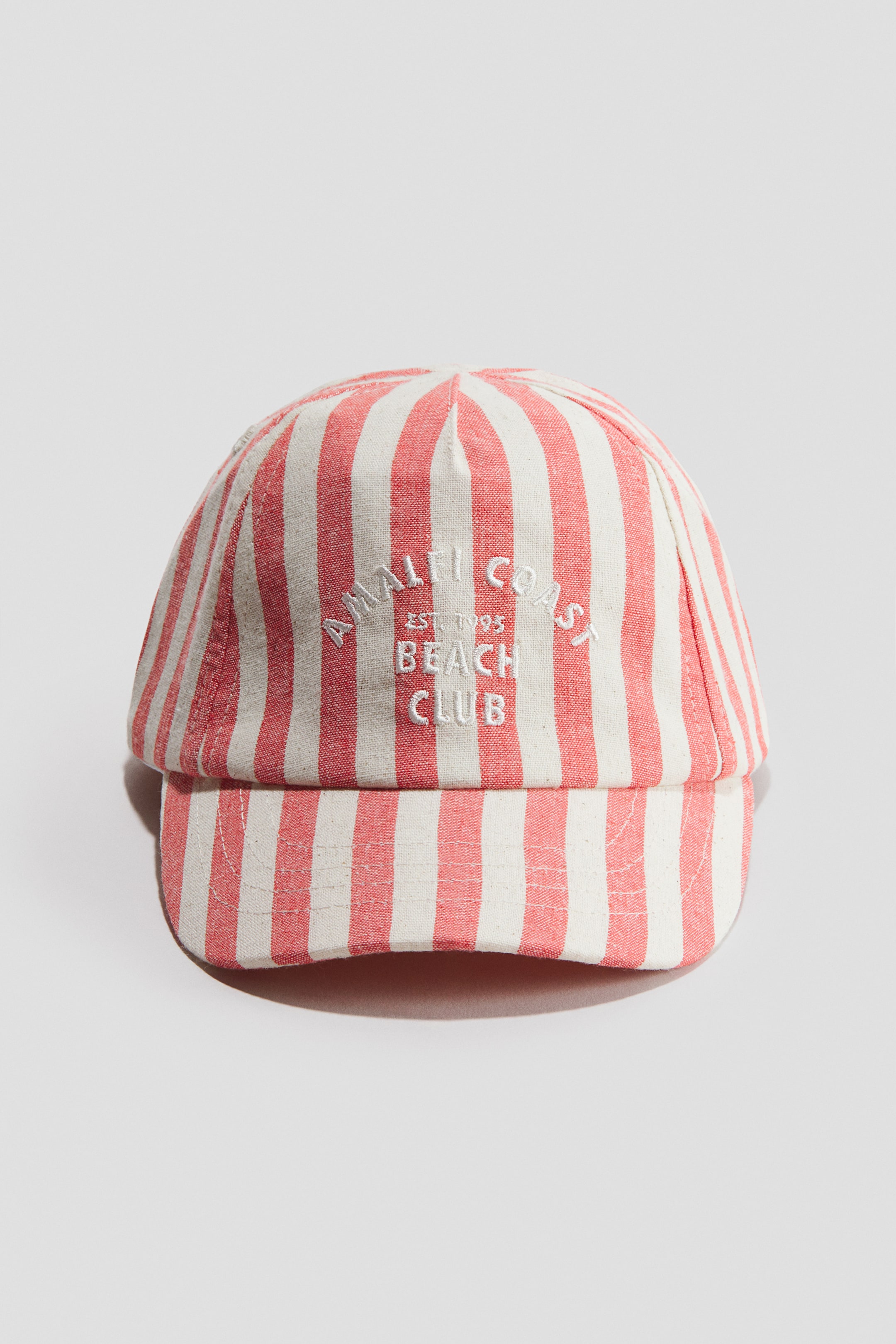 View larger image: A front-facing cap showcases a red and off-white vertical striped cotton weave. White embroidered text, reading 'AMALFI COAST EST. 1995 BEACH CLUB', is prominently displayed on its front.
