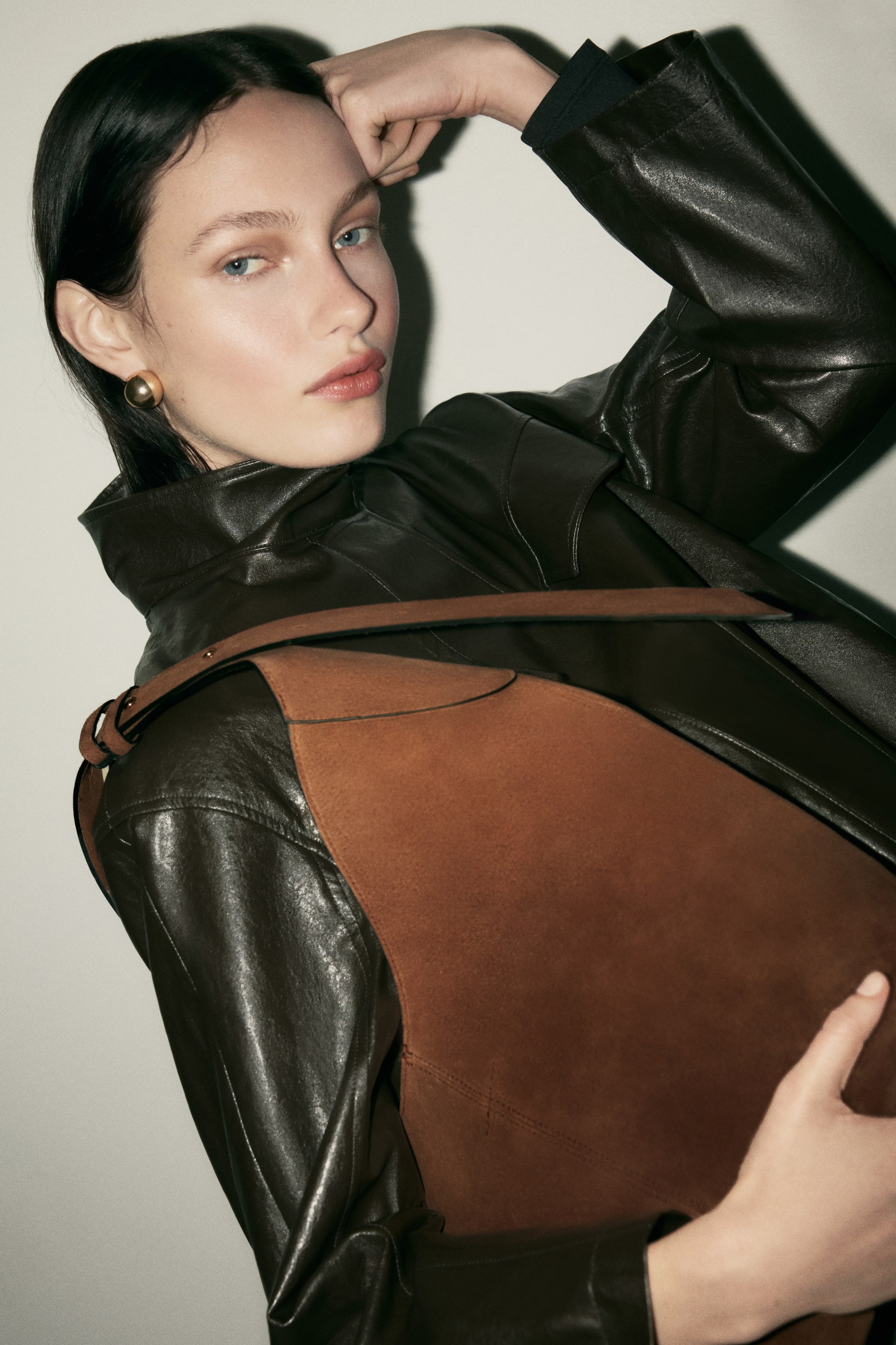 View larger image: A brown unstructured shoulder bag, crafted from a soft, textured material, is draped over the model's dark leather jacket. Its slender, dark strap rests on the model's shoulder, while the model holds the bag's body with one hand.