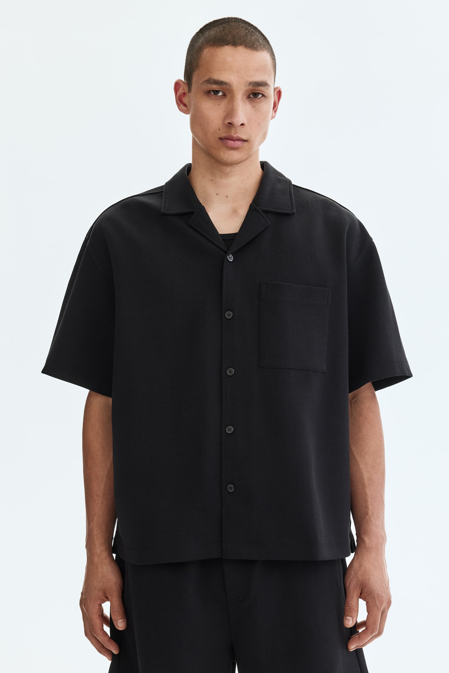 A black short-sleeved shirt featuring a camp collar and a button-front fastening with matching black buttons. It displays a single patch pocket on the left chest, a straight hem with side slits, and a black undershirt visible beneath its open collar. - 1