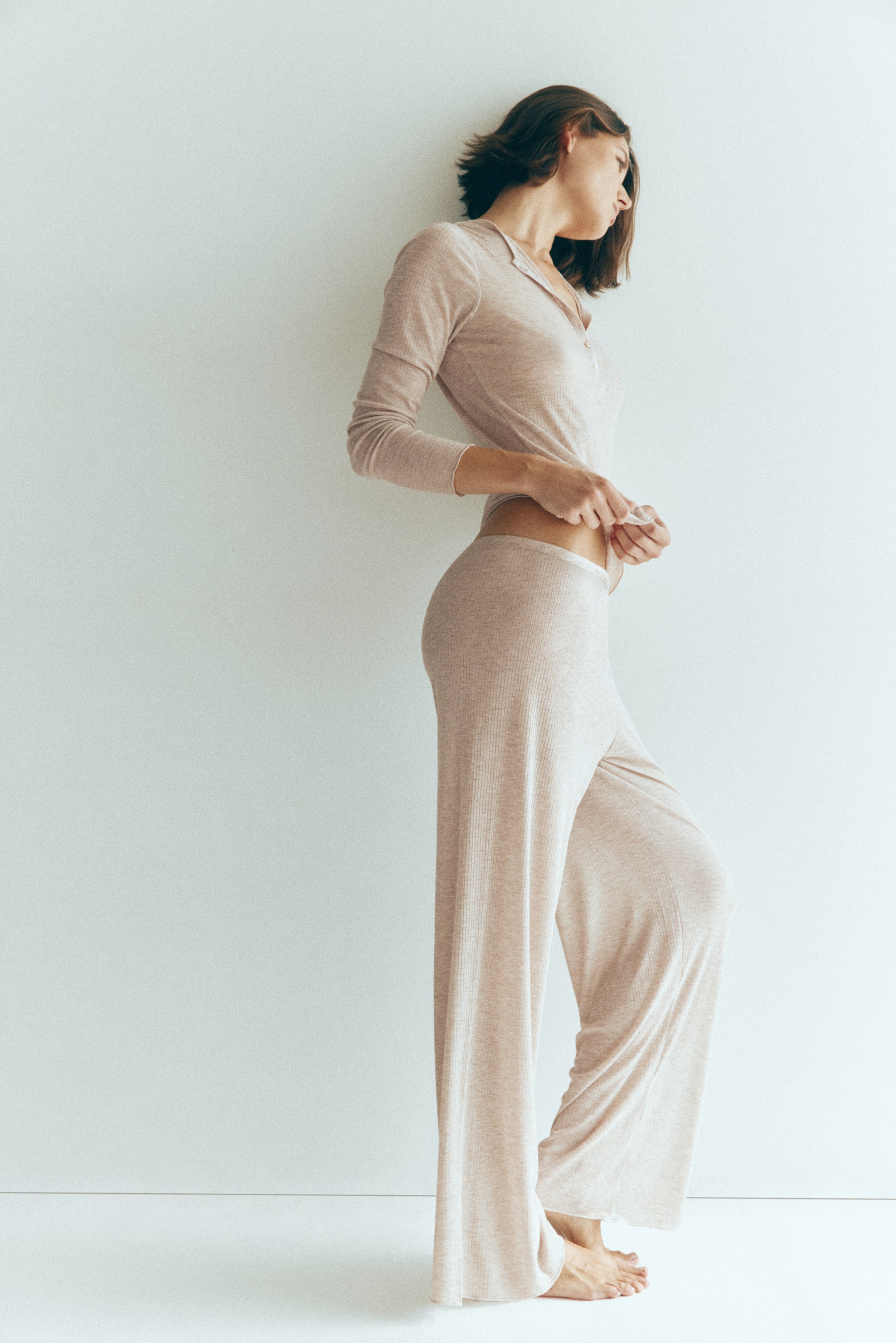 Visualizza immagine più grande: La modella indossa un completo pigiama coordinato in jersey a costine color beige chiaro, composto da una maglia a maniche lunghe con abbottonatura frontale e pantaloni a gamba ampia con orli rifiniti a punto overlock. La modella è in piedi di profilo e tiene il bordo inferiore della maglia.