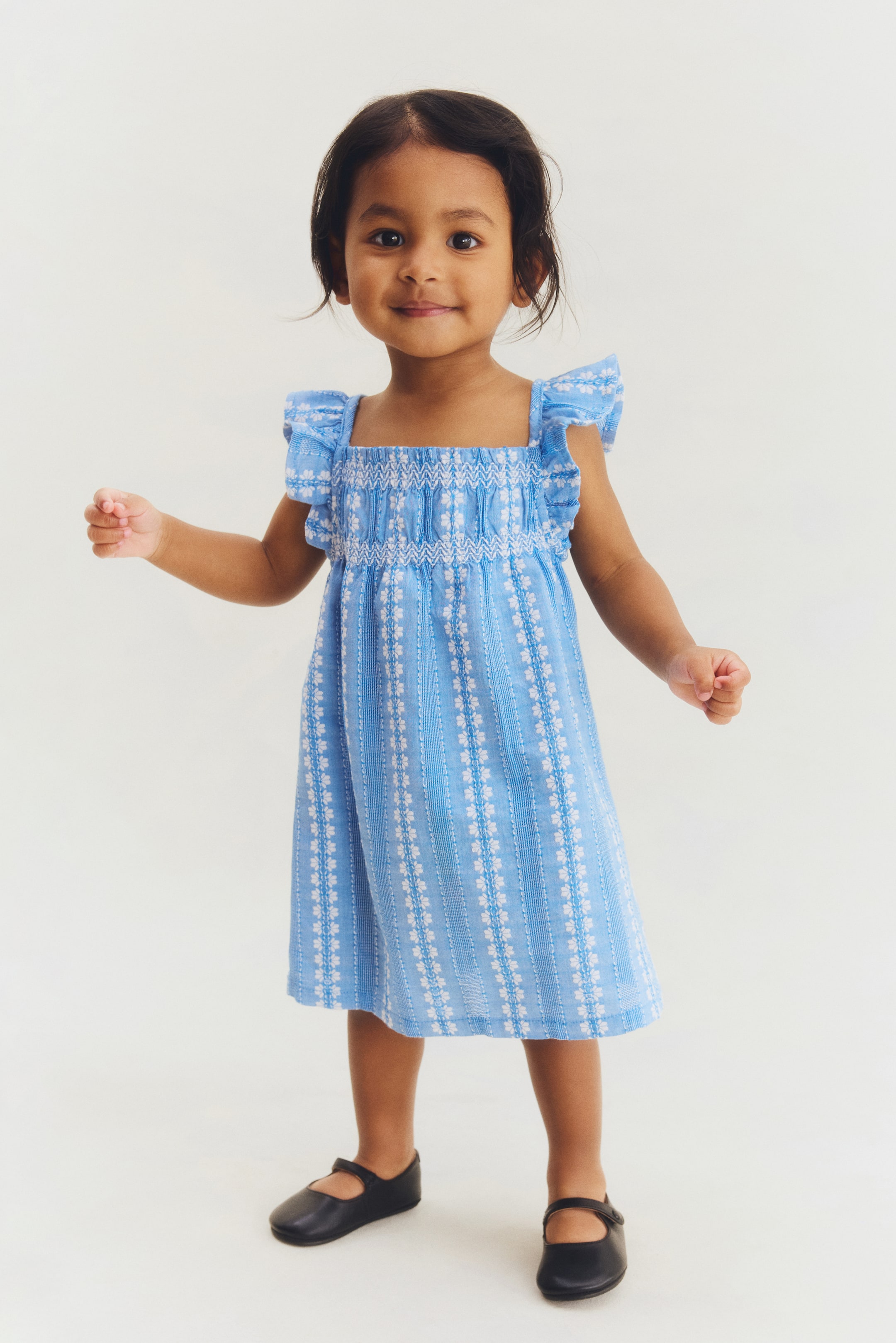 Größeres Bild ansehen: Ein hellblaues Kinderkleid mit einem quadratischen Ausschnitt und gerüschten Kappärmeln. Das Oberteil ist mit weißem Garn gesmokt, und der Rock zeigt vertikale Reihen von weißen Blumenmustern, die sich mit strukturierten Streifen abwechseln und bis zur Knielänge fließen.