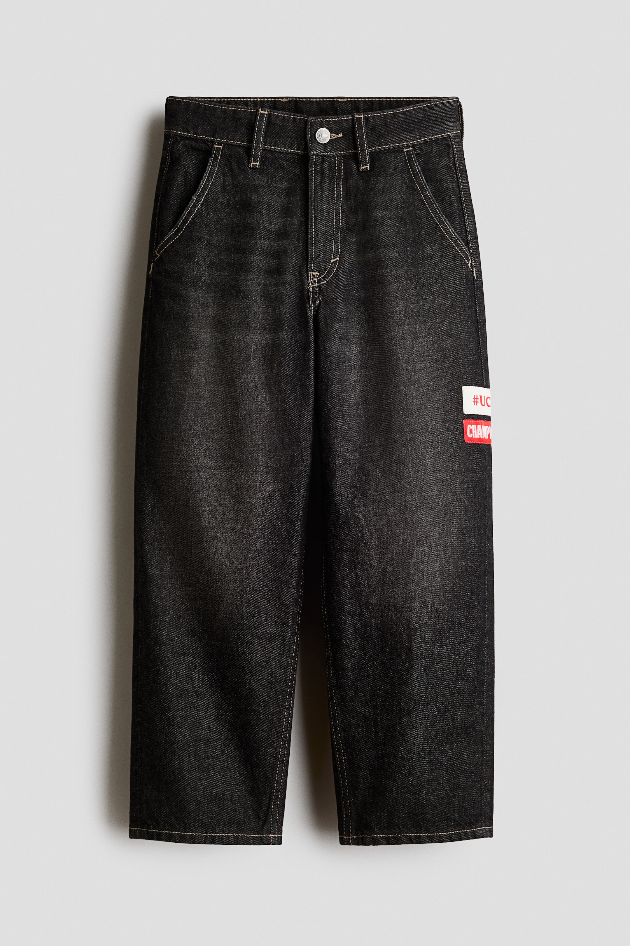 Grotere afbeelding bekijken: Zwarte denim broek met een gewassen afwerking en contrasterende lichte stiksels over het hele ontwerp. Voorzien van een sluiting met knoop en rits aan de voorkant, schuine steekzakken en een rechthoekige witte en rode patch met 'CHAMP'-tekst op het rechterbovenbeen.