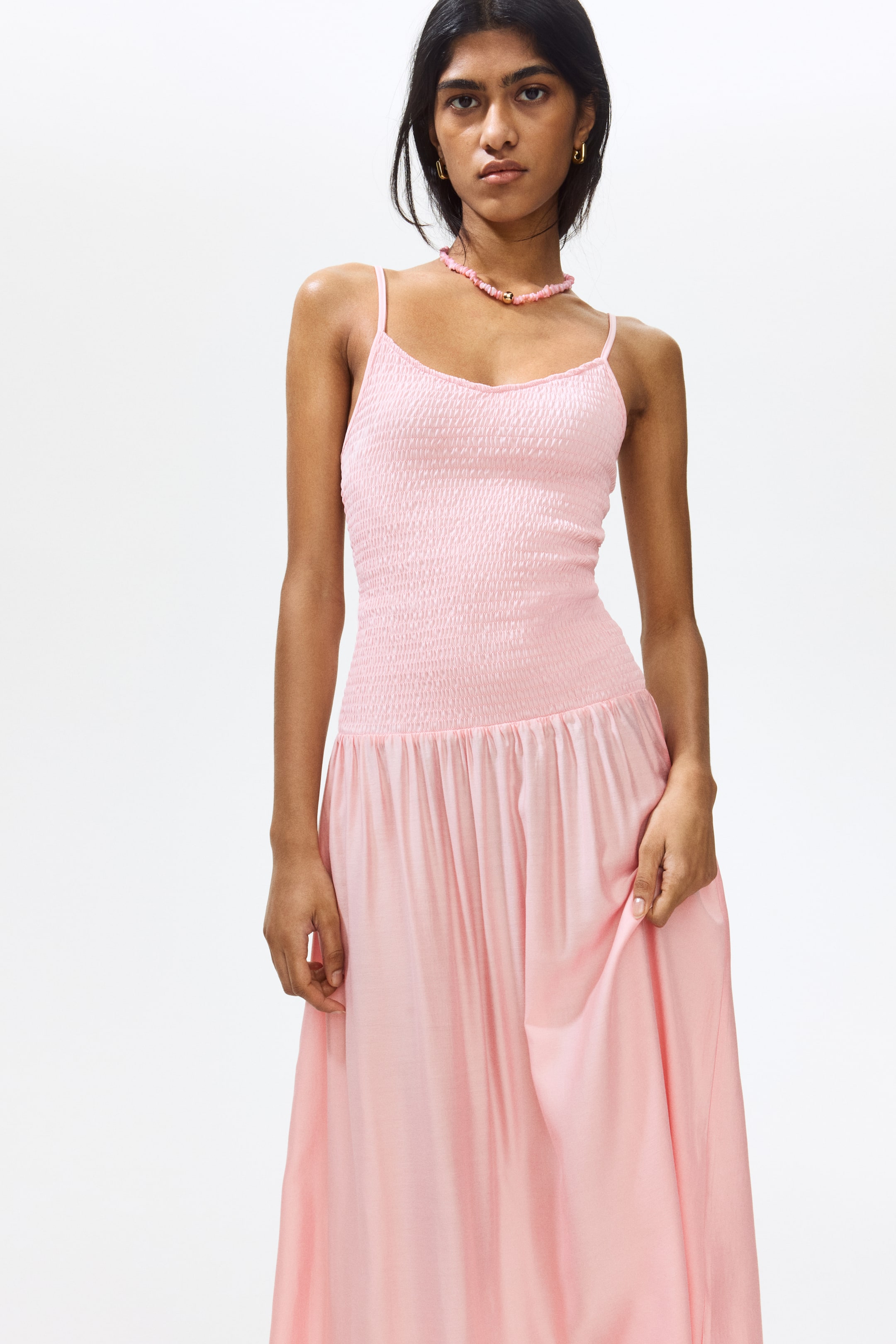 View larger image: A pastel pink maxi dress showcasing a V-neckline and thin spaghetti straps. The bodice is gathered, giving a textured, snug fit at the top, while the skirt elegantly flows from a gathered waistline to a maxi length.