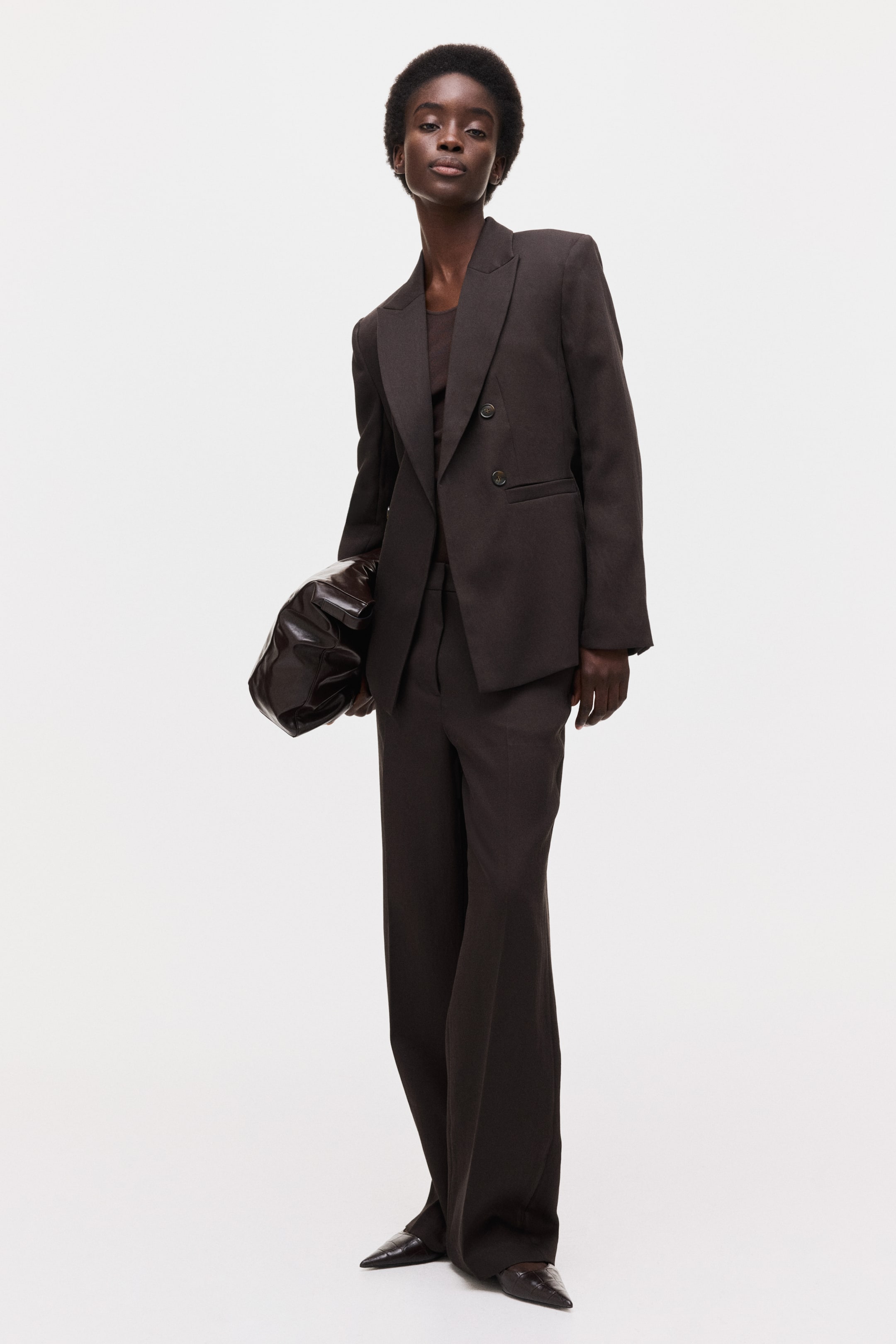 拡大画像を表示: ダークブラウンのダブルブレストブレザー(ノッチドラペル)を同色のトップスの上に羽織り、ワイドパンツと合わせています。スーツはやや左向きに配置され、構築的な肩のラインと流れるようなシルエットが際立っています。