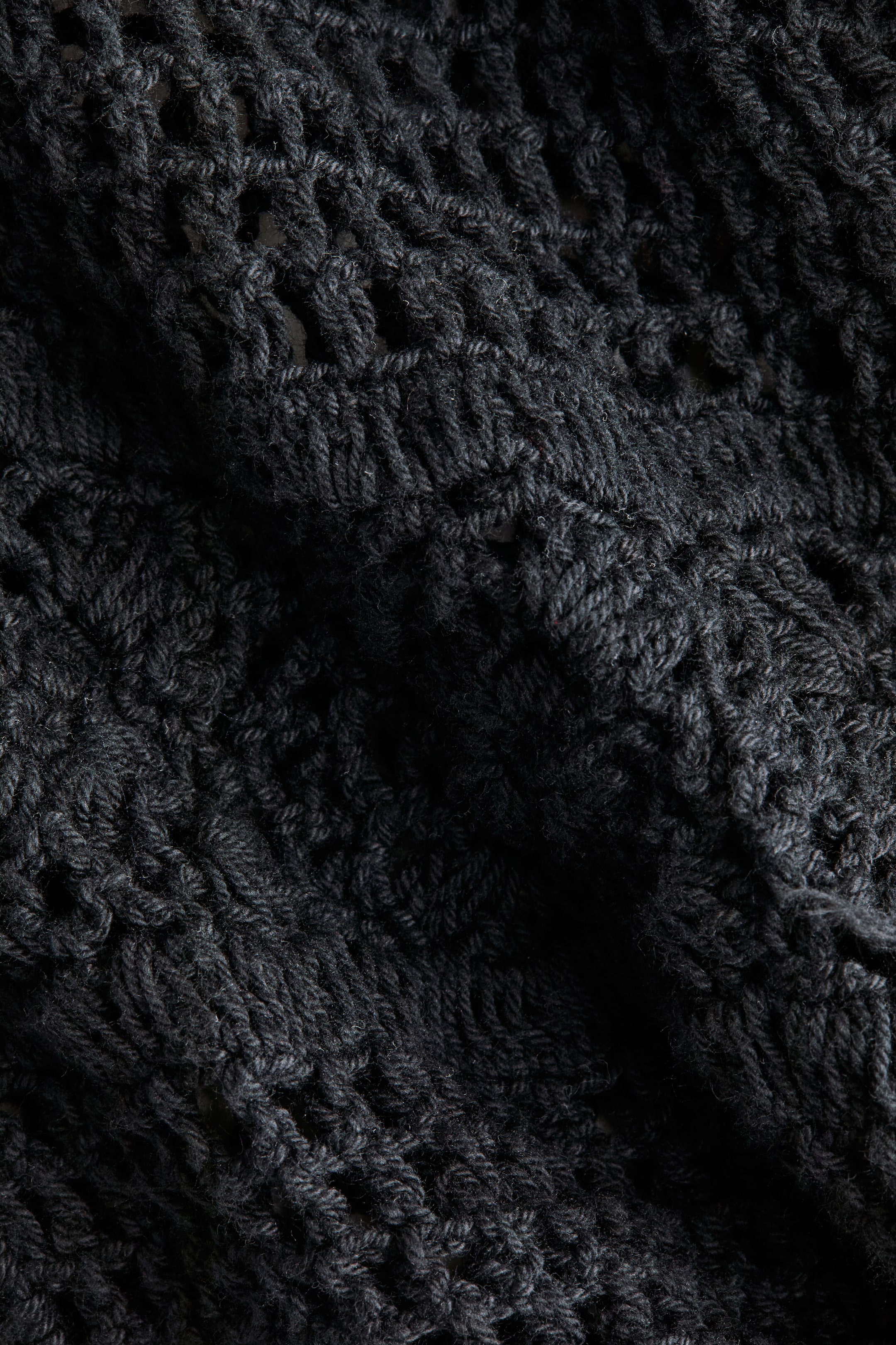 Agrandir l'image: Un tissu tricoté gris foncé affiche un motif texturé, avec un maillage ajouré associé à des points plus denses et en relief. Le tissu tombe avec fluidité.