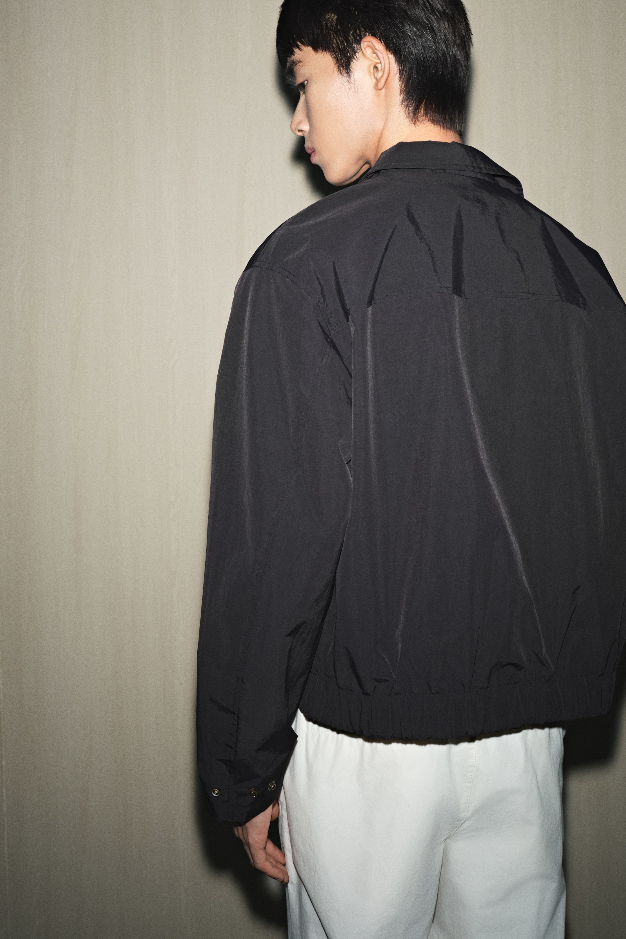 拡大画像を表示: 黒の軽量ジャケットが後ろ左の角度から見せられており、そのリラックスしたフィット感が強調されています。それは、尖った襟、ギャザー入りの伸縮性のある裾、調節可能なスナップカフスを特徴としており、上部の背中に水平の縫い目のディテールが見えます。