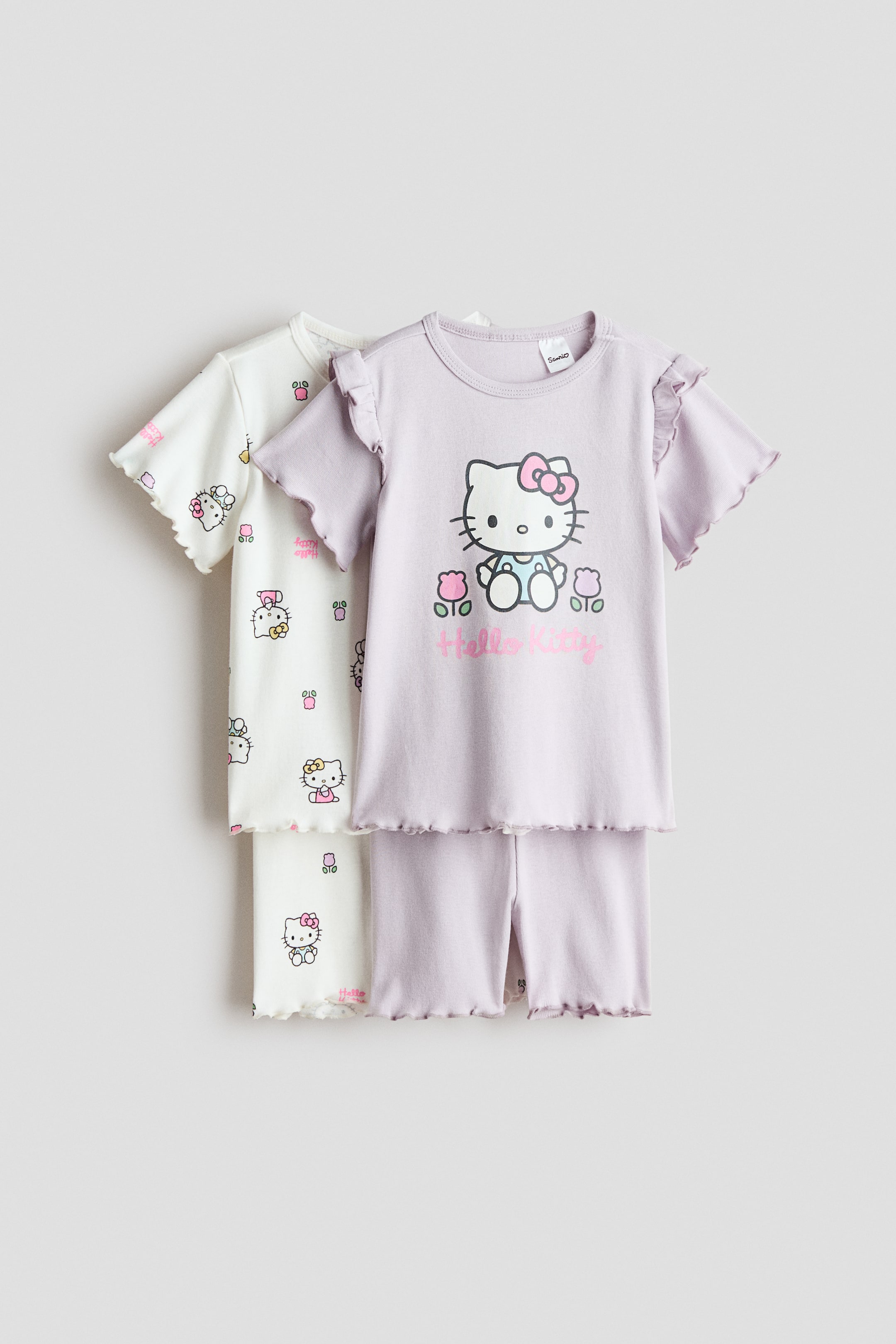 View larger image: Two sets of children's nightwear are positioned facing the front, both featuring lettuce-edge hems. The light purple set showcases a short-sleeved top with ruffle details on the sleeves and a prominent Hello Kitty graphic on the front, paired with matching shorts. The white set, layered in the background, features a short-sleeved top and shorts adorned with an all-over Hello Kitty pattern.