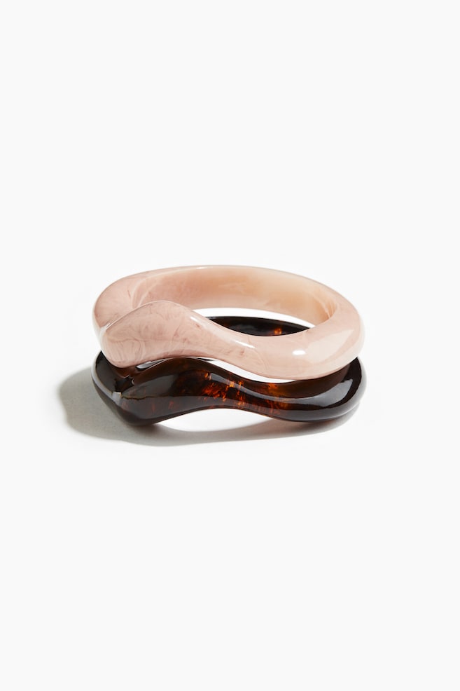 Two chunky, glossy plastic bangles, featuring an irregular, wavy shape, are resting one on top of the other. The top bangle is light pink with a marbled pattern, and the bottom bangle is dark brown with a tortoiseshell pattern. - 1