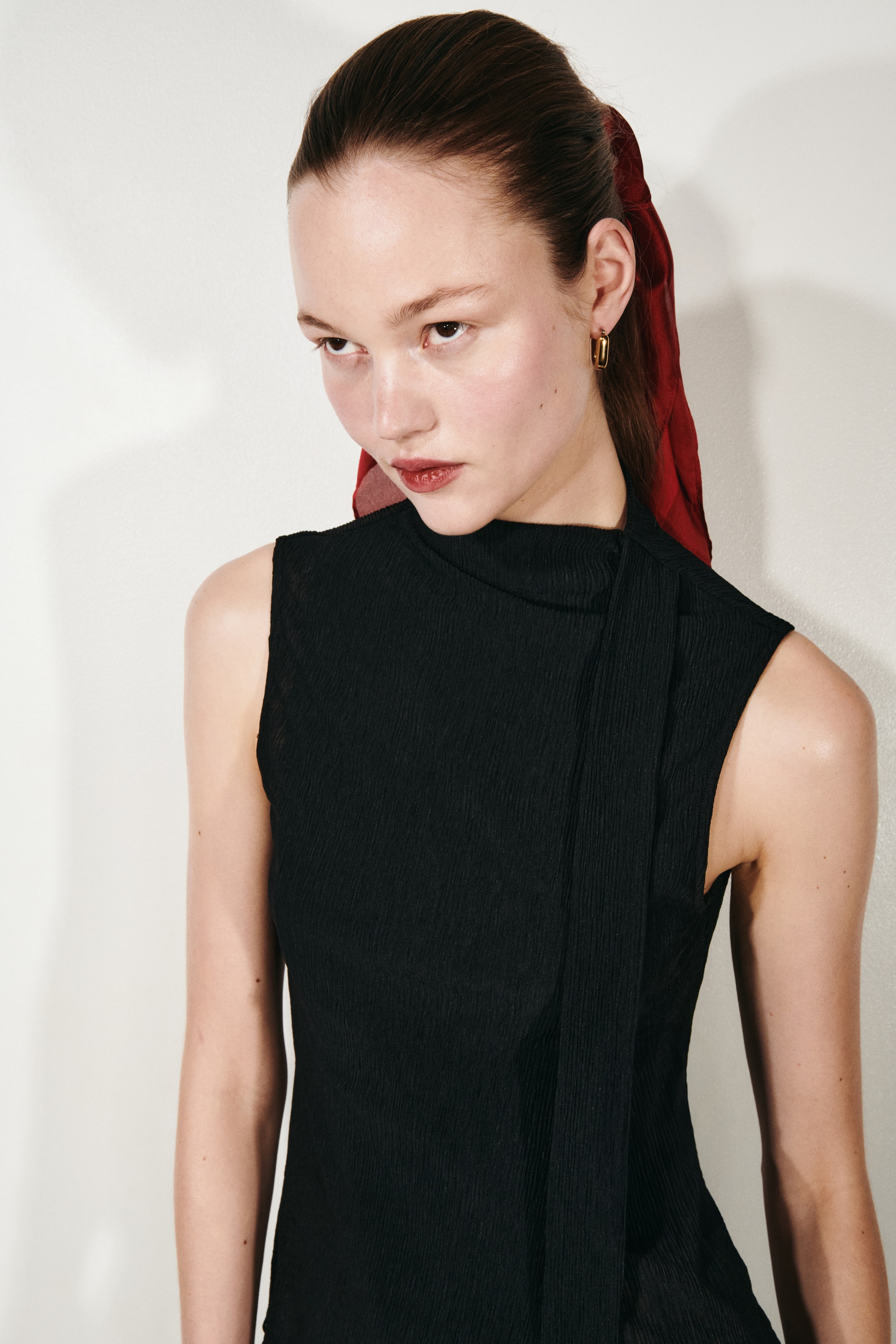 View larger image: A black sleeveless top showcasing a high mock neck and an asymmetrical panel draping from the right shoulder across the front, all crafted from a finely pleated fabric.