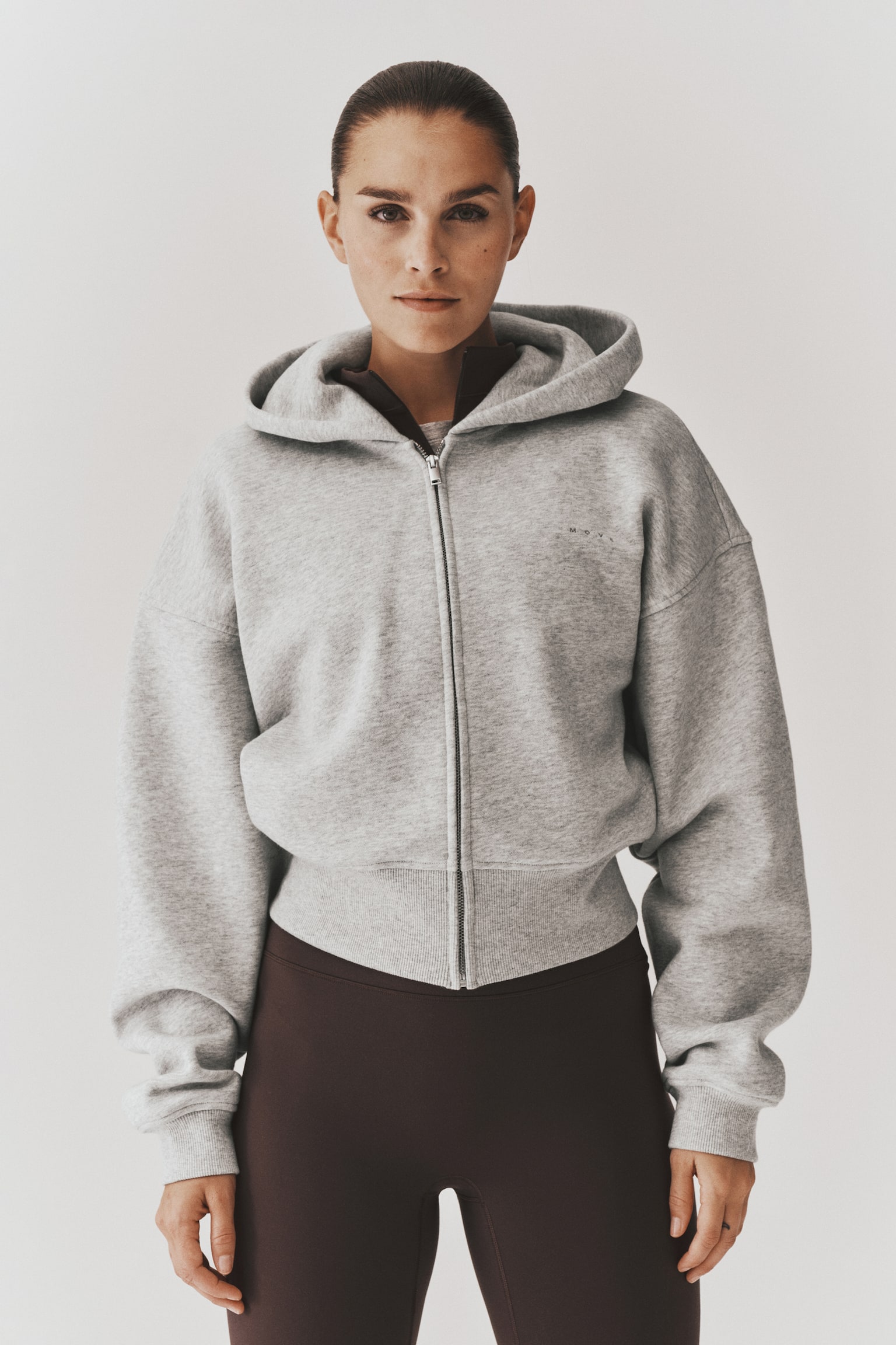 A light grey zip-up cropped hoodie is shown from the front, featuring a relaxed fit with dropped shoulders, a hood, and a front zip fastening. The hoodie is designed with a wide ribbed hem and ribbed cuffs. - 1
