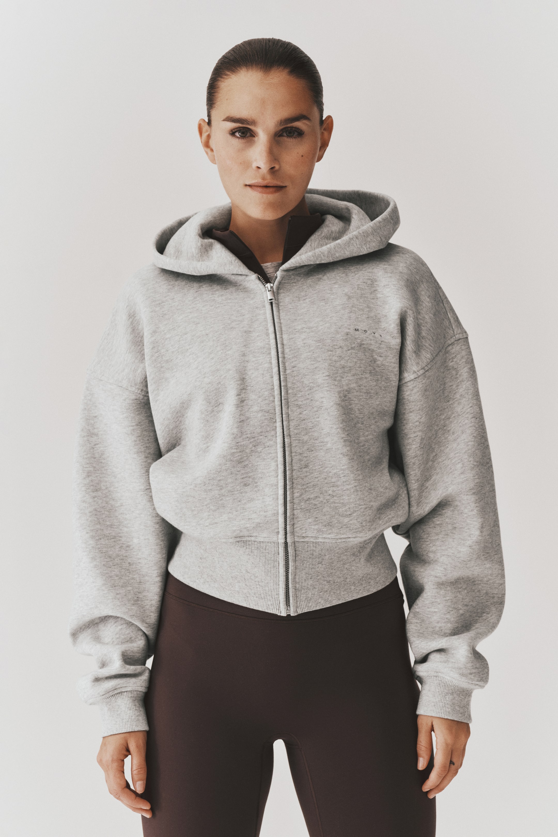 View larger image: A light grey cropped sports hoodie with a double-layered hood, dropped shoulders, and a front zip is being worn by the model. The loose-fitting hoodie features ribbed cuffs and a wide ribbed hem, and is layered over a dark brown high-collar top and high-waisted leggings.