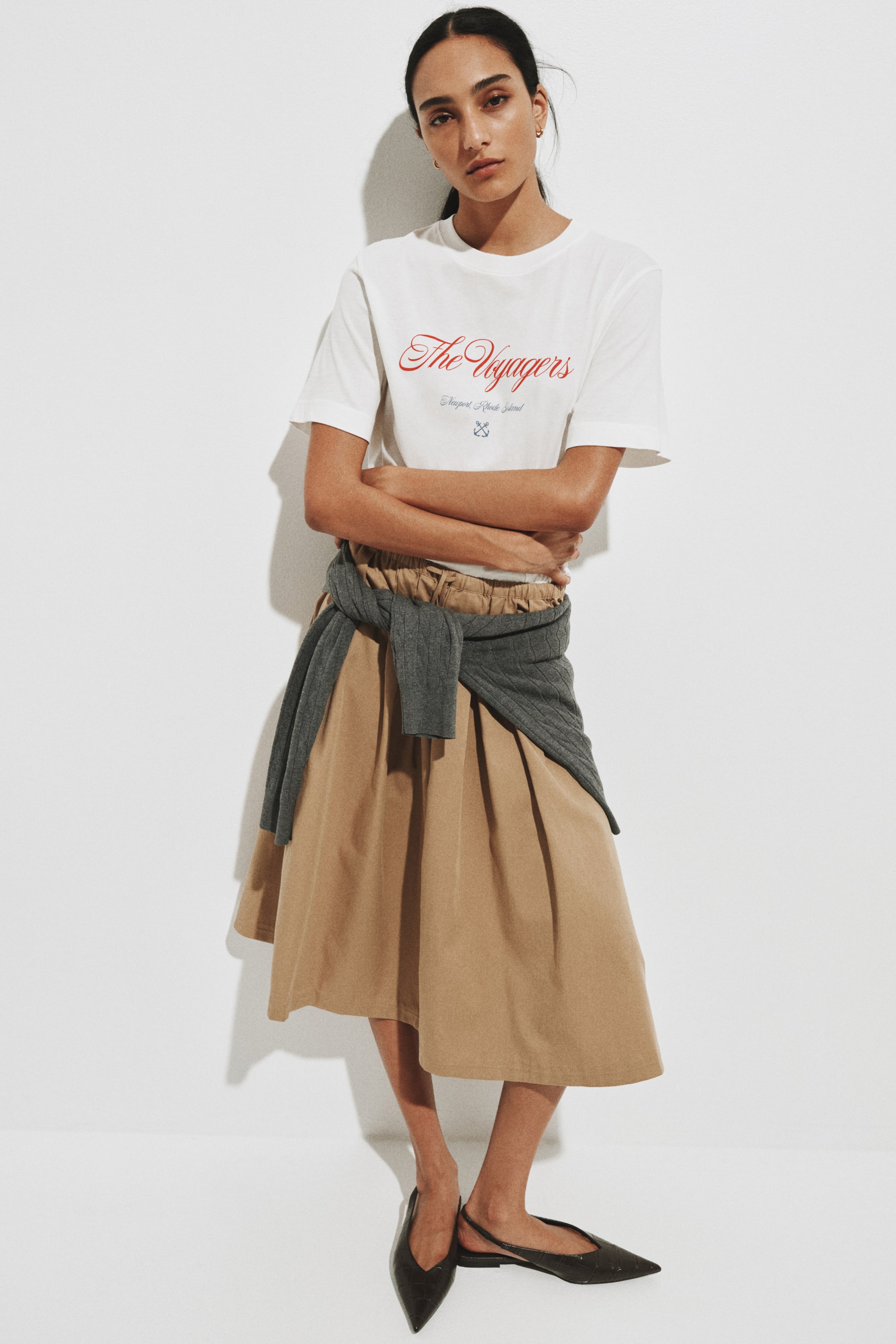 查看大圖: 白色圓領短袖T恤以紅色草寫字體印有 "The Voyagers"，下方印有 "Newport, Rhode Island" 字樣及藍色交叉槳圖案。短袖長度至上臂中段，衣襬紮入裙內穿著。