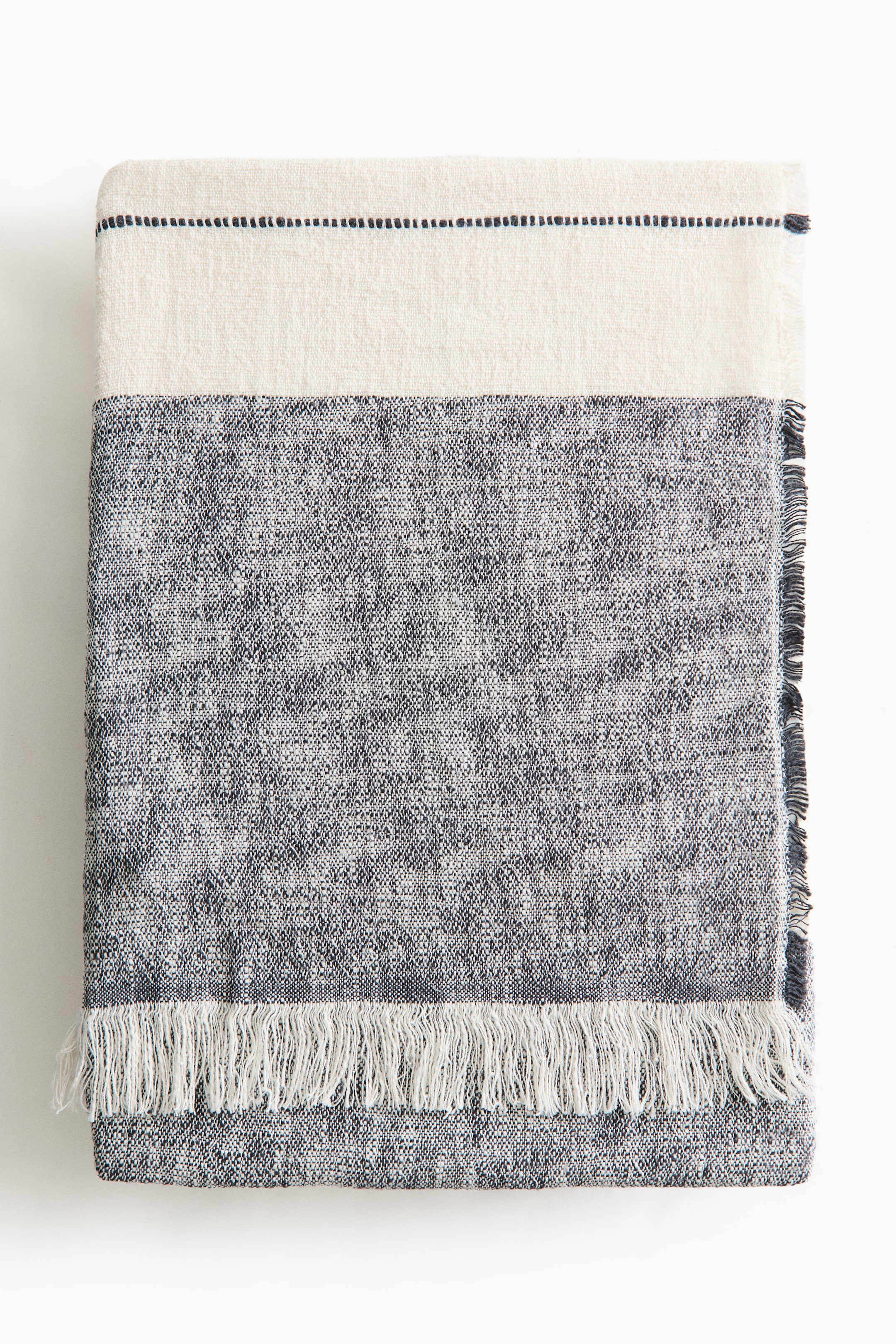 View larger image: A blanket in a soft weave made from a linen and cotton blend features a striped pattern with frayed edges and fringes along the short sides. The pattern displays a wide cream band at the top, separated by a thin dotted black line from a mottled grey and white section, with long white fringes at the bottom edge.
