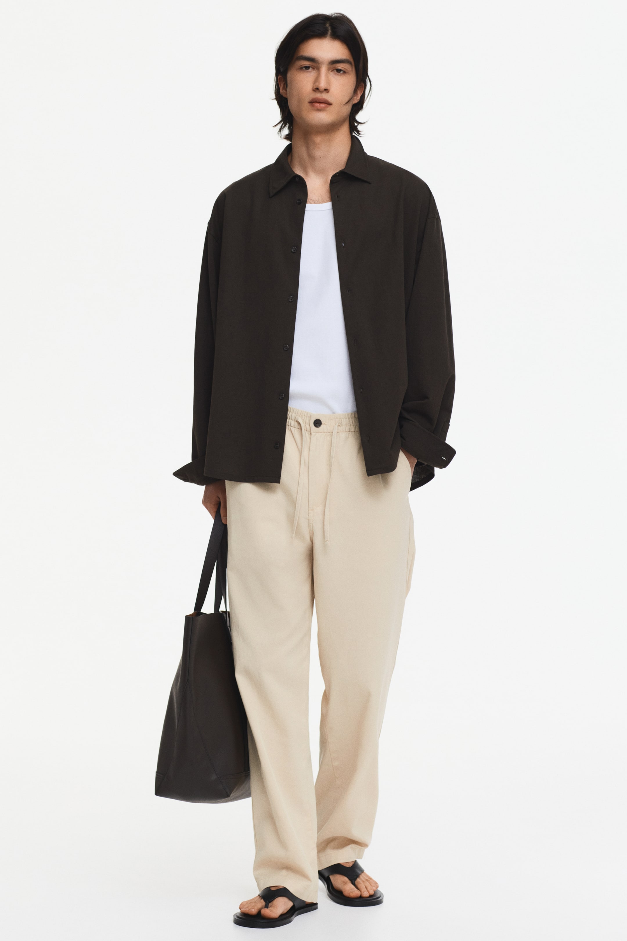 View larger image: Light beige, full-length trousers feature an elasticated drawstring waist with the drawstring tied. They present a relaxed, wide-leg fit, with hands resting in the side pockets.