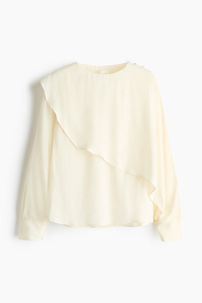 A cream blouse is shown from the front, featuring a round neckline and an asymmetrical draped overlay across the front. It is designed with long sleeves, buttoned cuffs, and small buttons detailing the right shoulder. - 1