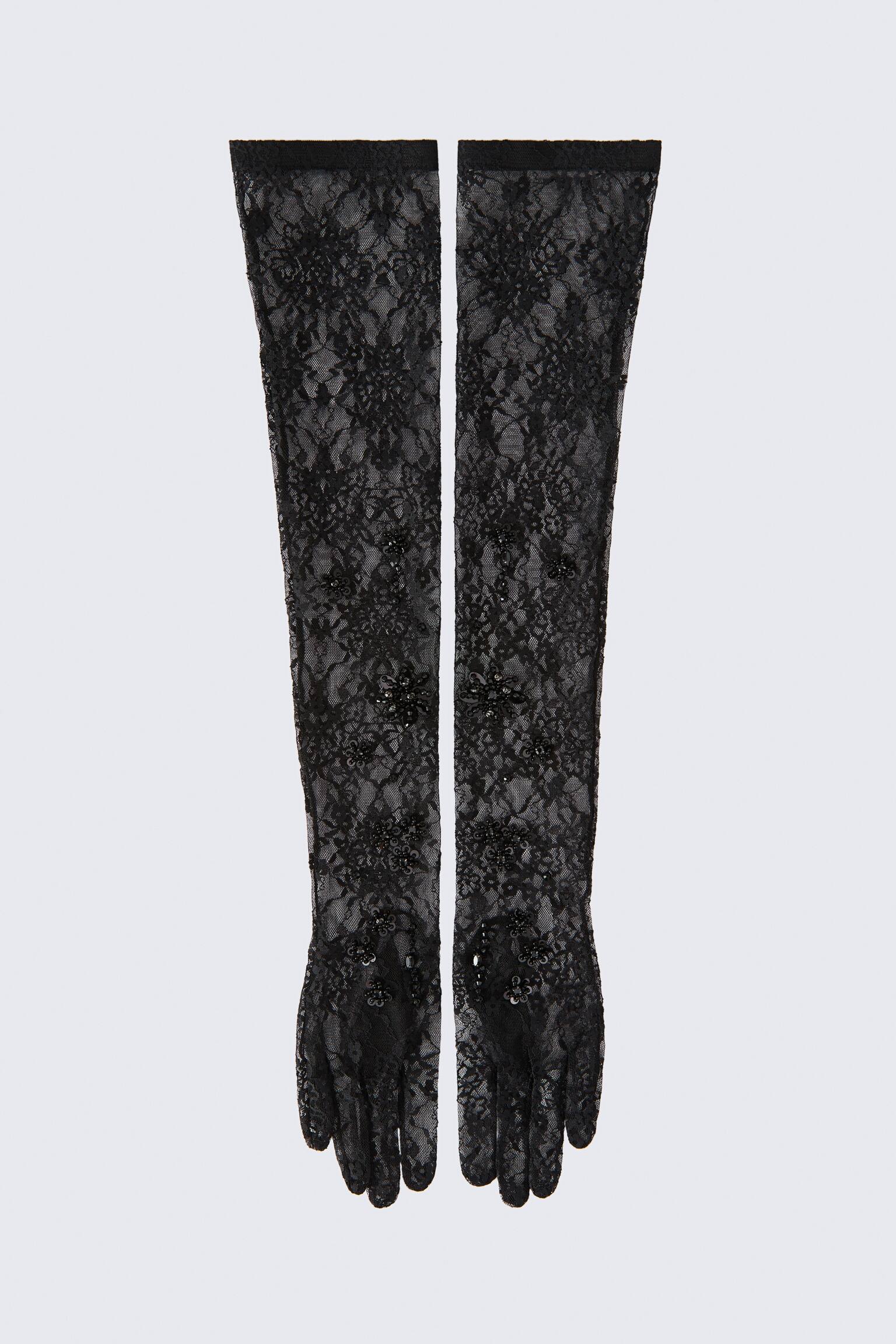 A pair of long, black stretch-lace gloves is lying flat, displaying a sheer floral pattern adorned with matching black beads and sequins. The unlined, opera-length gloves feature narrow elasticated bands around the top openings.