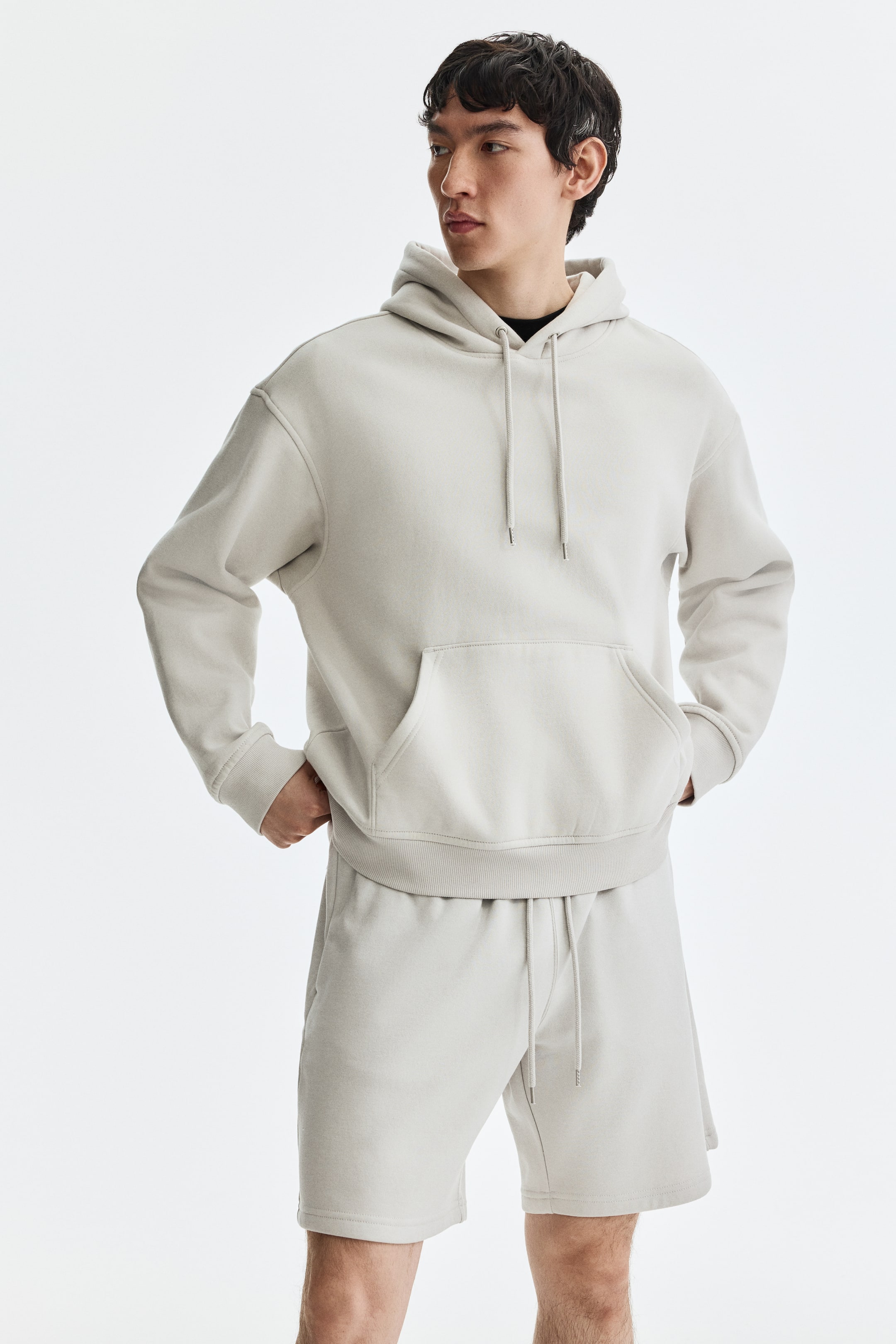 View larger image: A light beige hoodie and matching shorts are showcased from a front-facing, slightly angled view. The hoodie features a drawstring hood, a front kangaroo pocket, ribbed cuffs, and a ribbed hem, while the shorts have a drawstring waist.
