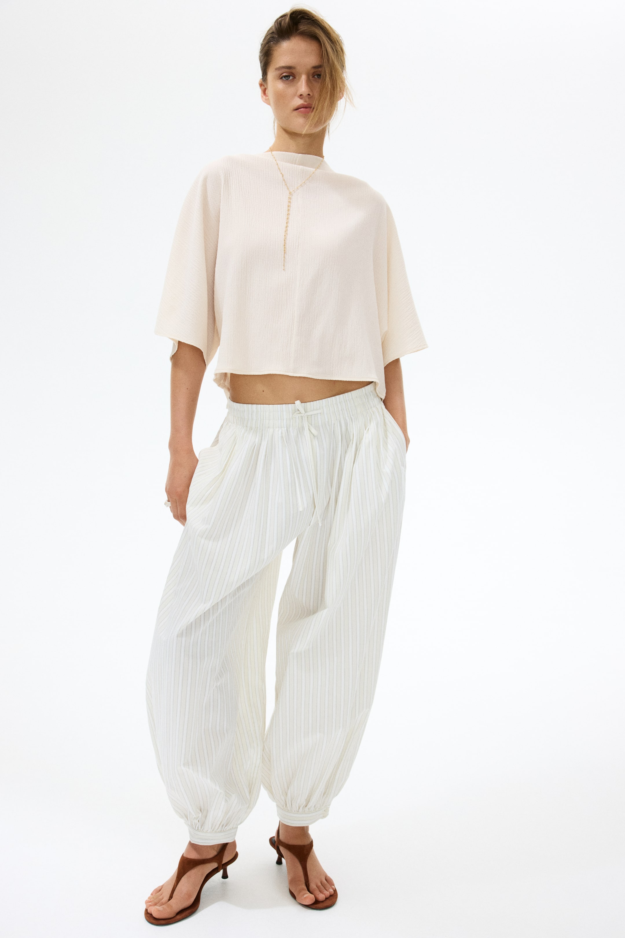 Voir une image plus grande: Un haut court texturé beige clair, avec un col montant et de larges manches courtes, est assorti à un pantalon ballon à fines rayures blanches. Le pantalon présente une taille élastiquée avec cordon de serrage et des chevilles froncées, tandis qu'un long collier doré est drapé sur le haut et des sandales à talons de style tong marron sont visibles.