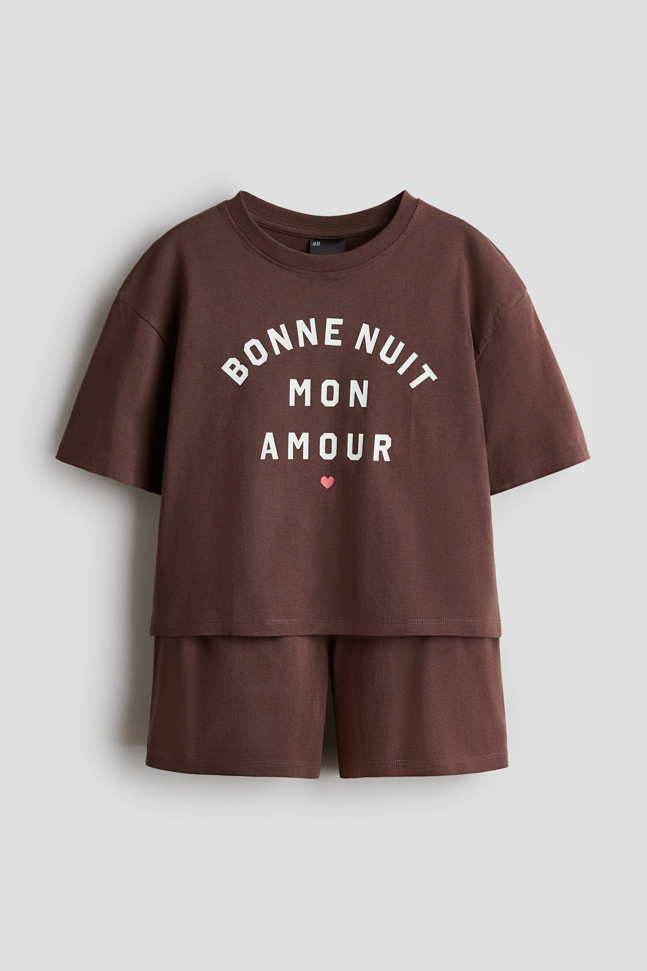 View larger image: A brown soft cotton jersey pyjama set is showcased from the front, highlighting a top with a round neckline, dropped shoulders, and short sleeves. The top displays "BONNE NUIT MON AMOUR" in white text with a pink heart on the front, and the matching shorts feature an elasticated waist.