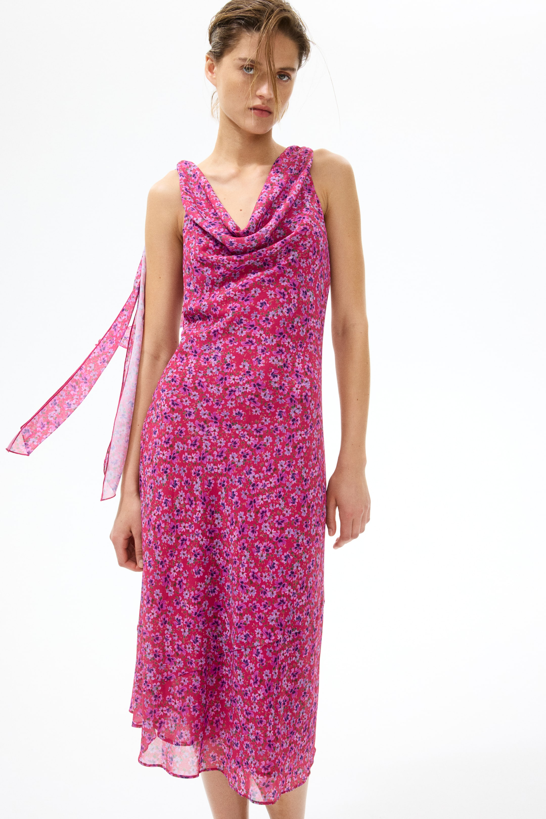 View larger image: A pink midi dress is showcased front-facing, highlighting a delicate floral pattern in shades of pink and purple. It features a cowl neckline and a sleeveless design, with two long fabric ties draping from the left shoulder.