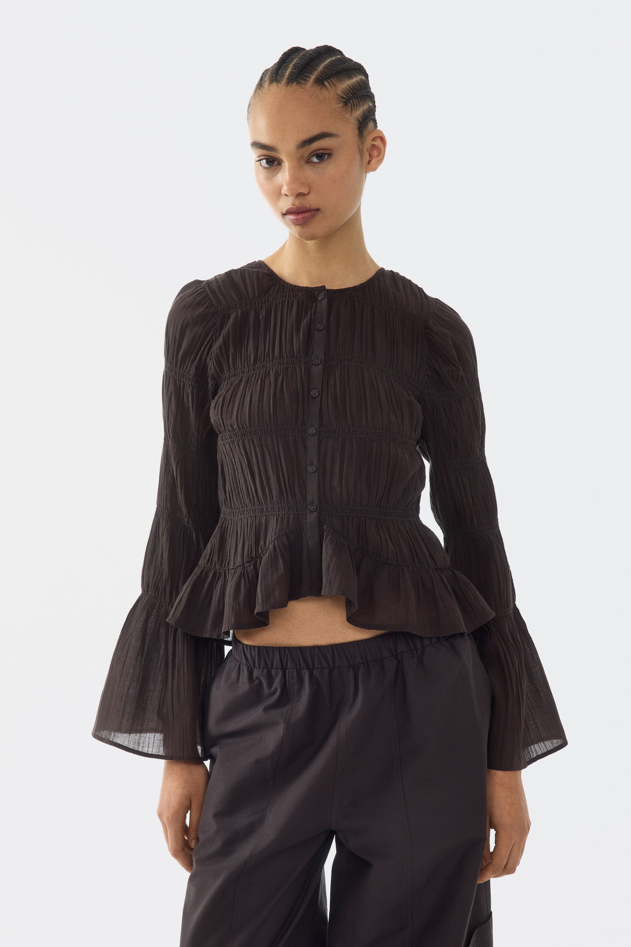 View larger image: The dark brown peplum top features a round neckline, a full button placket down the front, and long, shirred bell sleeves, all crafted from a textured, gathered fabric throughout, with a ruffled hem that ends above the waist.