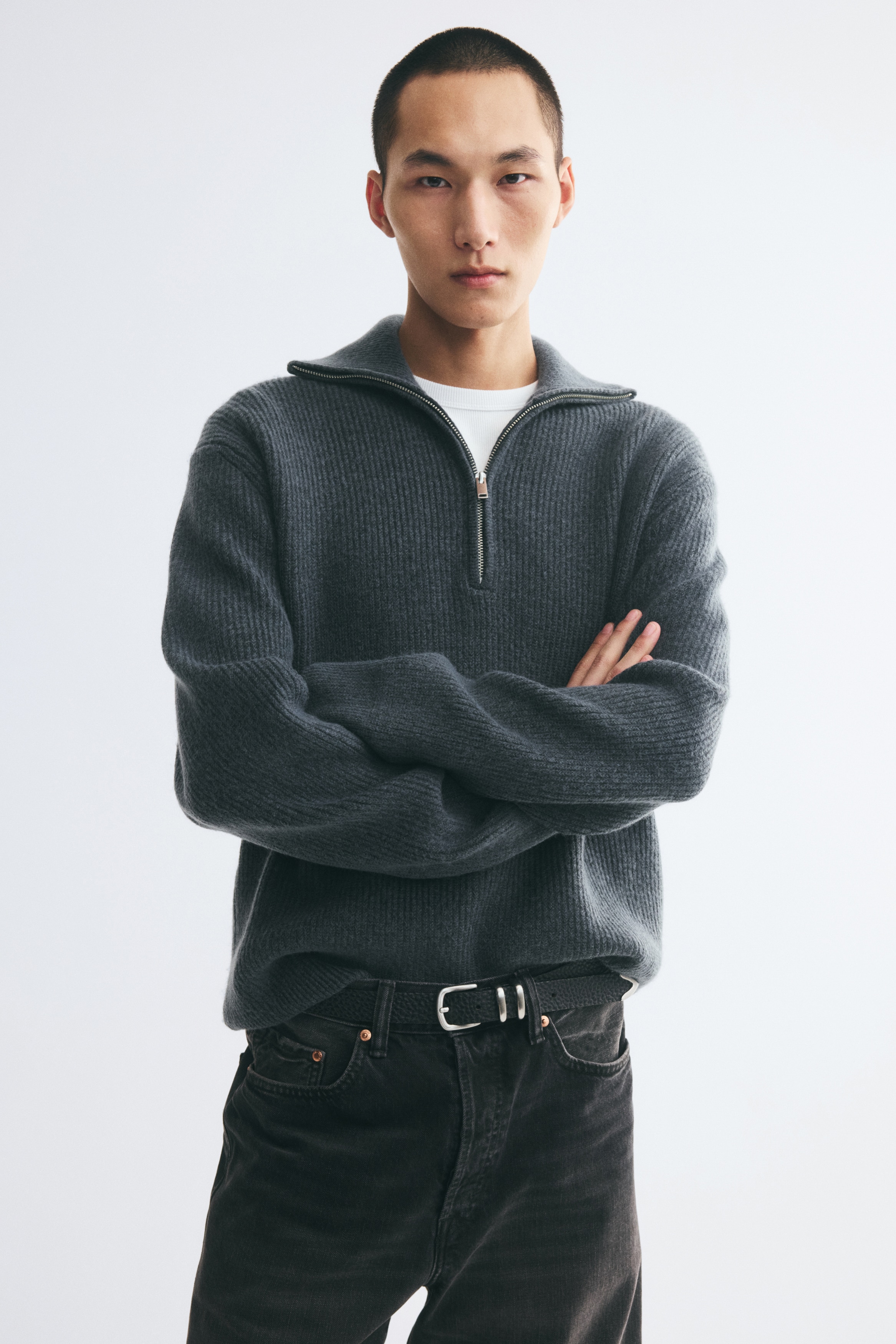 A dark grey ribbed-knit half-zip sweater is shown from the front, featuring a high funnel-neck collar with a silver-tone zipper. The sweater showcases an all-over vertical ribbed texture, complete with long sleeves, ribbed cuffs, and a straight hem.