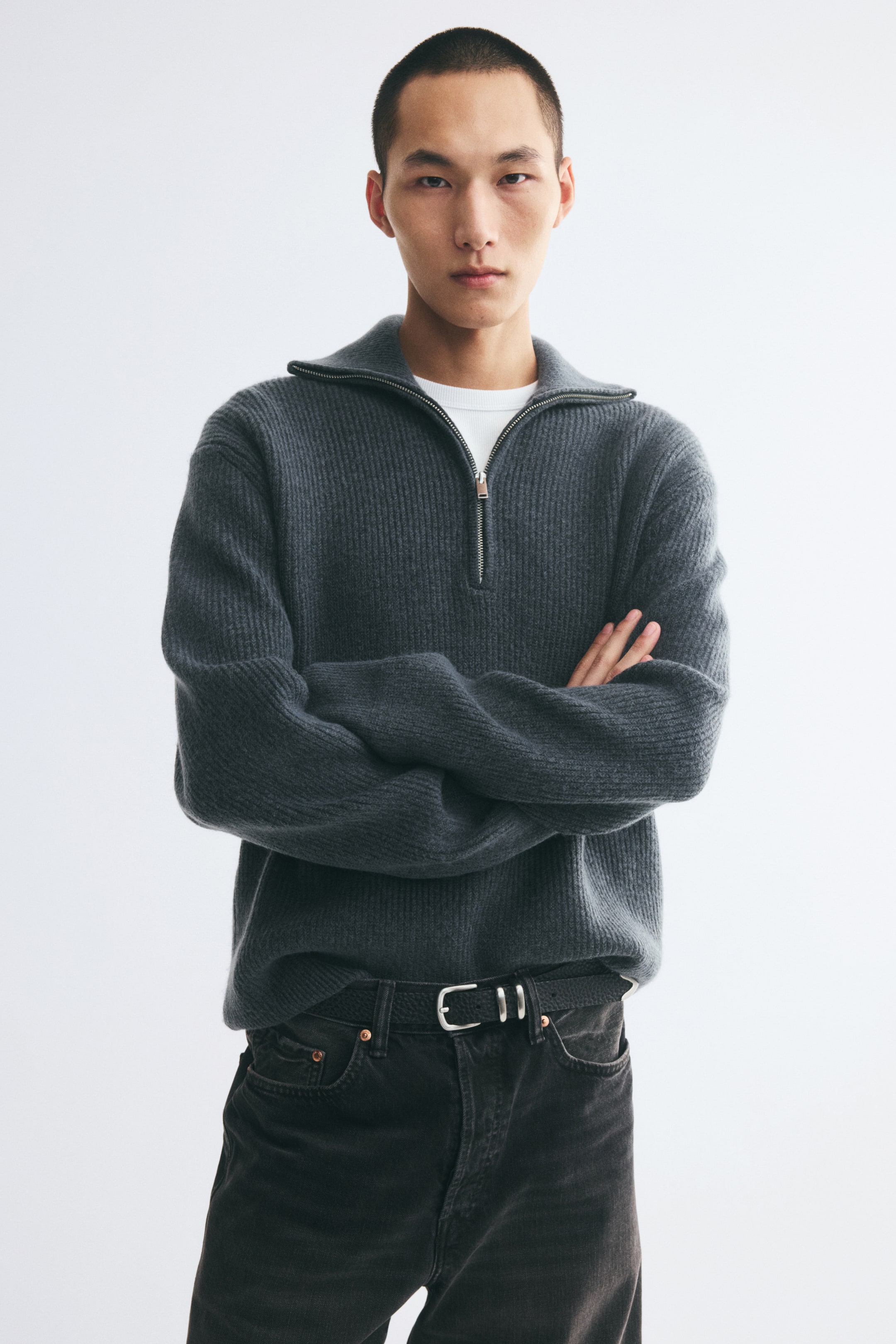 View larger image: A dark grey, rib-knit sweater with a half-zip collar is being worn by the model, who is standing with arms crossed. The relaxed-fit sweater is layered over a white crew-neck T-shirt and styled with black jeans.