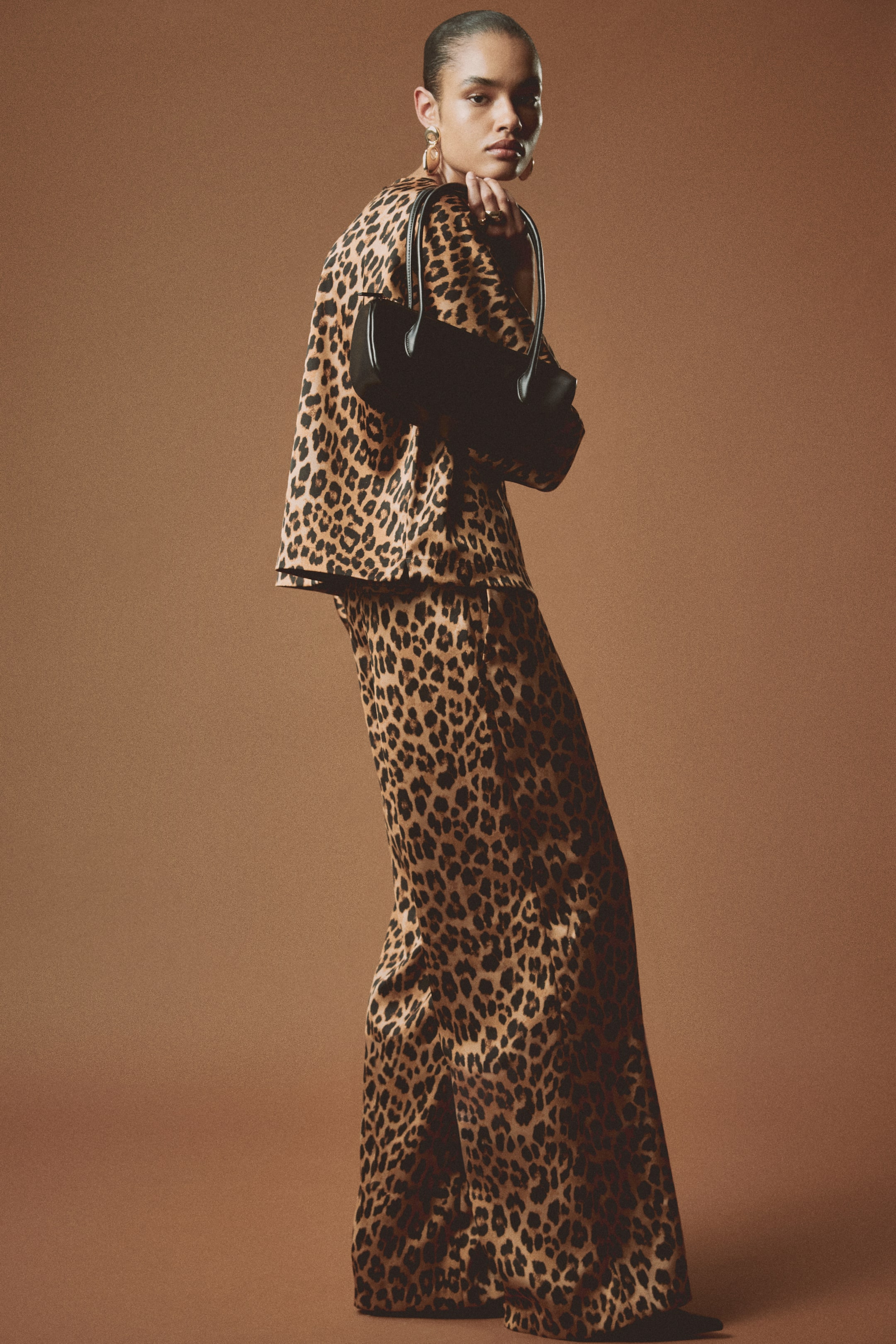 View larger image: A coordinating leopard print two-piece set, featuring a relaxed long-sleeve top and wide-leg trousers in a silky fabric, is being showcased. A small black cylindrical handbag with two top handles is being held, highlighting its smooth material from a front-facing perspective.