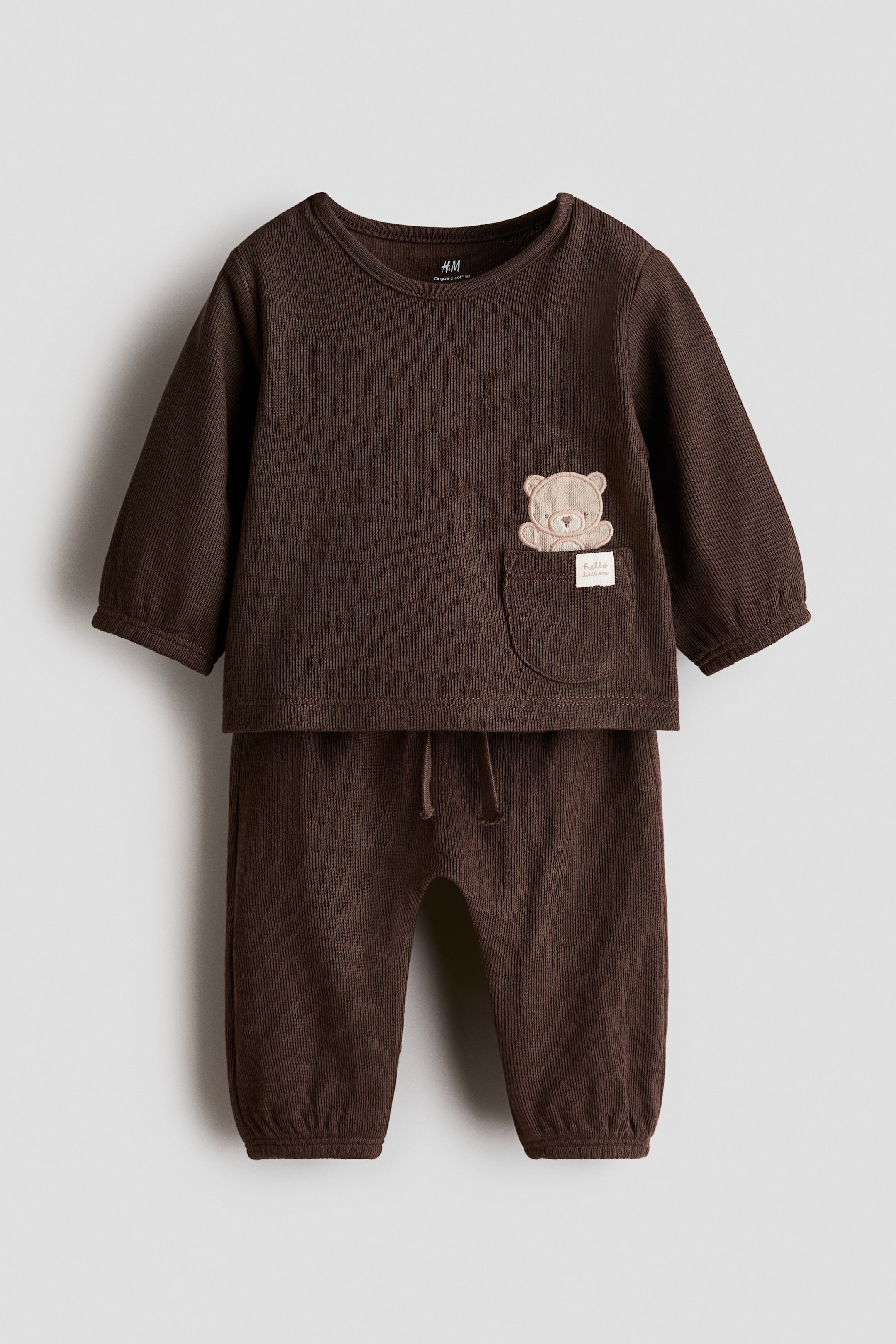 View larger image: A dark brown ribbed baby set is shown from the front, featuring a long-sleeve top with elasticated cuffs and a patch pocket with a small teddy bear applique. The matching pants feature an elasticated waistband with a decorative drawstring and elasticated cuffs.