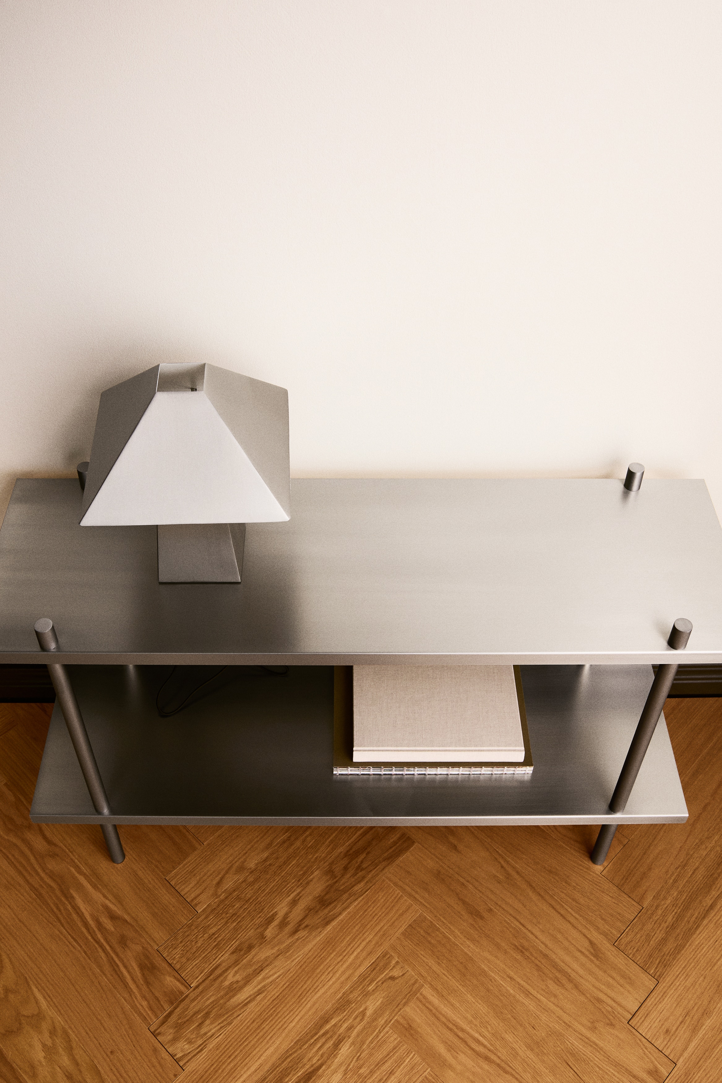 Una estanterÃ­a metÃ¡lica de dos niveles aparece de frente, con dos baldas planas y rectangulares con acabado plateado mate. Las baldas estÃ¡n sostenidas por cuatro postes metÃ¡licos cilÃ­ndricos que atraviesan las esquinas de cada nivel y se prolongan hacia abajo para formar las patas.
