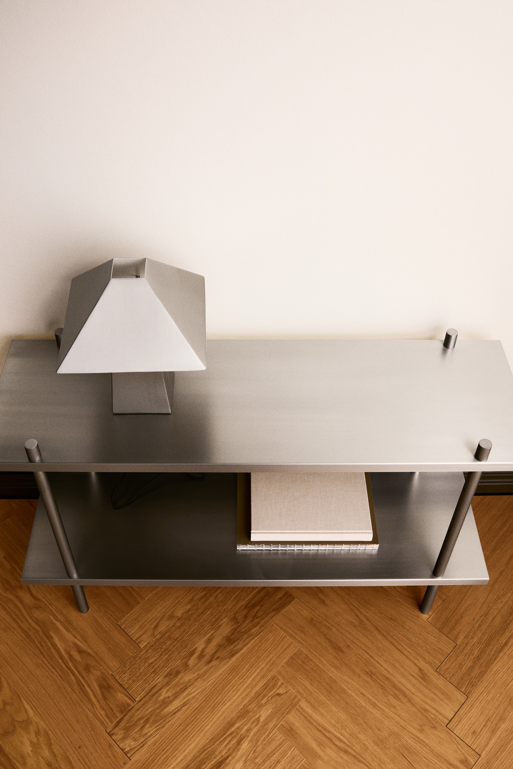 Ver imagen más grande: Se muestra una estantería de suelo de acero inoxidable con un elegante acabado cepillado, vista desde un ángulo frontal elevado. El diseño de dos niveles presenta cuatro postes cilíndricos en las esquinas, con una lámpara metálica geométrica sobre la balda superior y libros apoyados en la balda inferior.